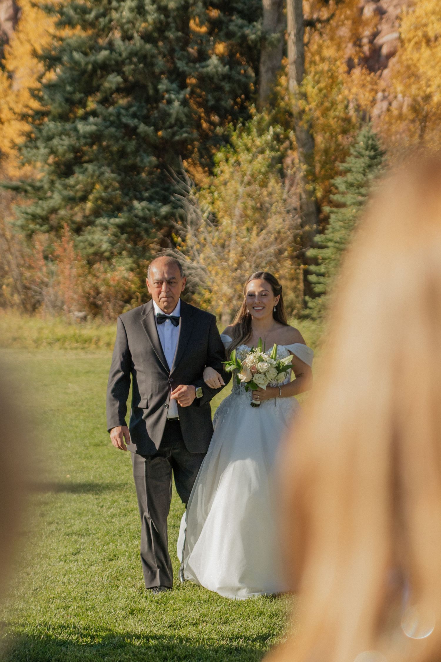 Outdoor wedding ceremony procession with autumn foliage in the background.
