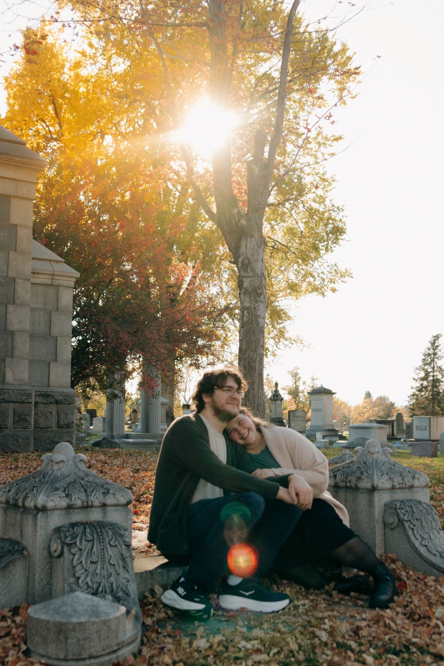 A couple sits together among fallen autumn leaves near stone architecture as golden sunlight filters through trees.