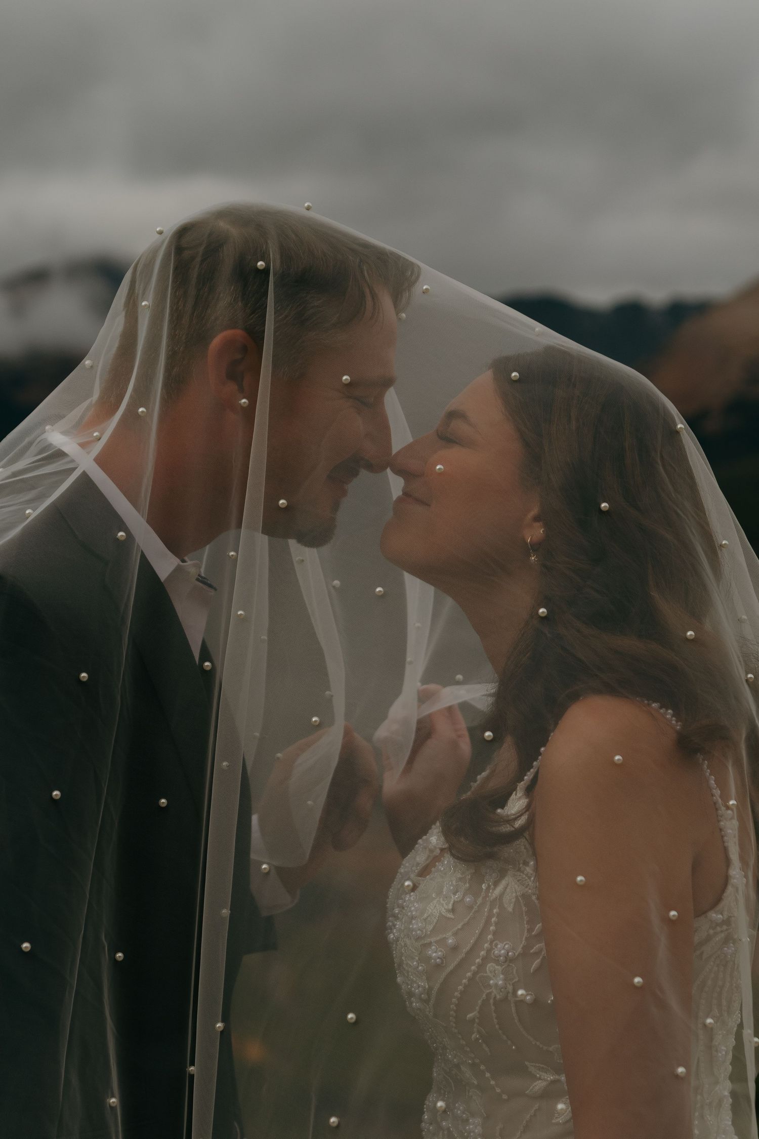 Romantic wedding couple shares an intimate moment under a delicate veil against a moody outdoor backdrop.