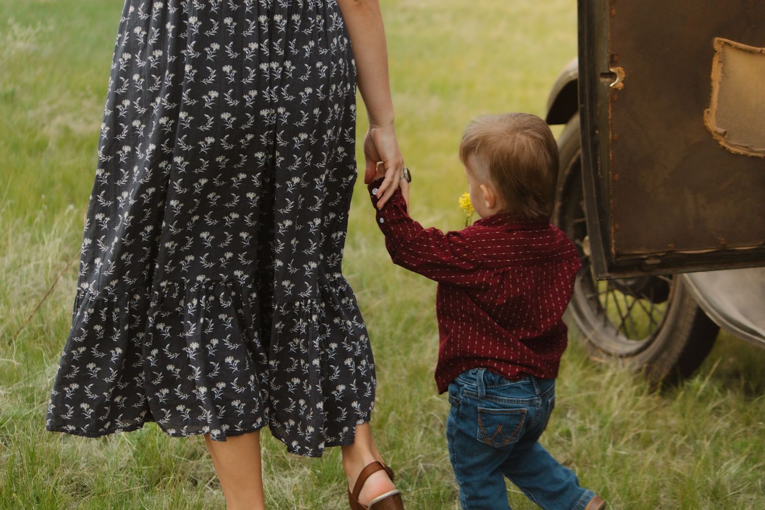 Person in a floral dress holding hands with a child next to an antique car.
