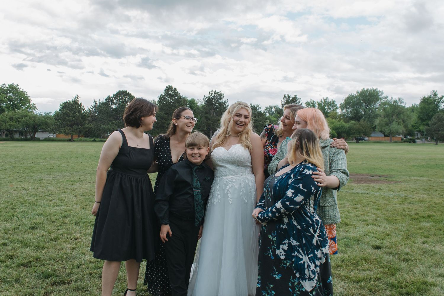 Wedding party group photo on grass field with cloudy sky backdrop.