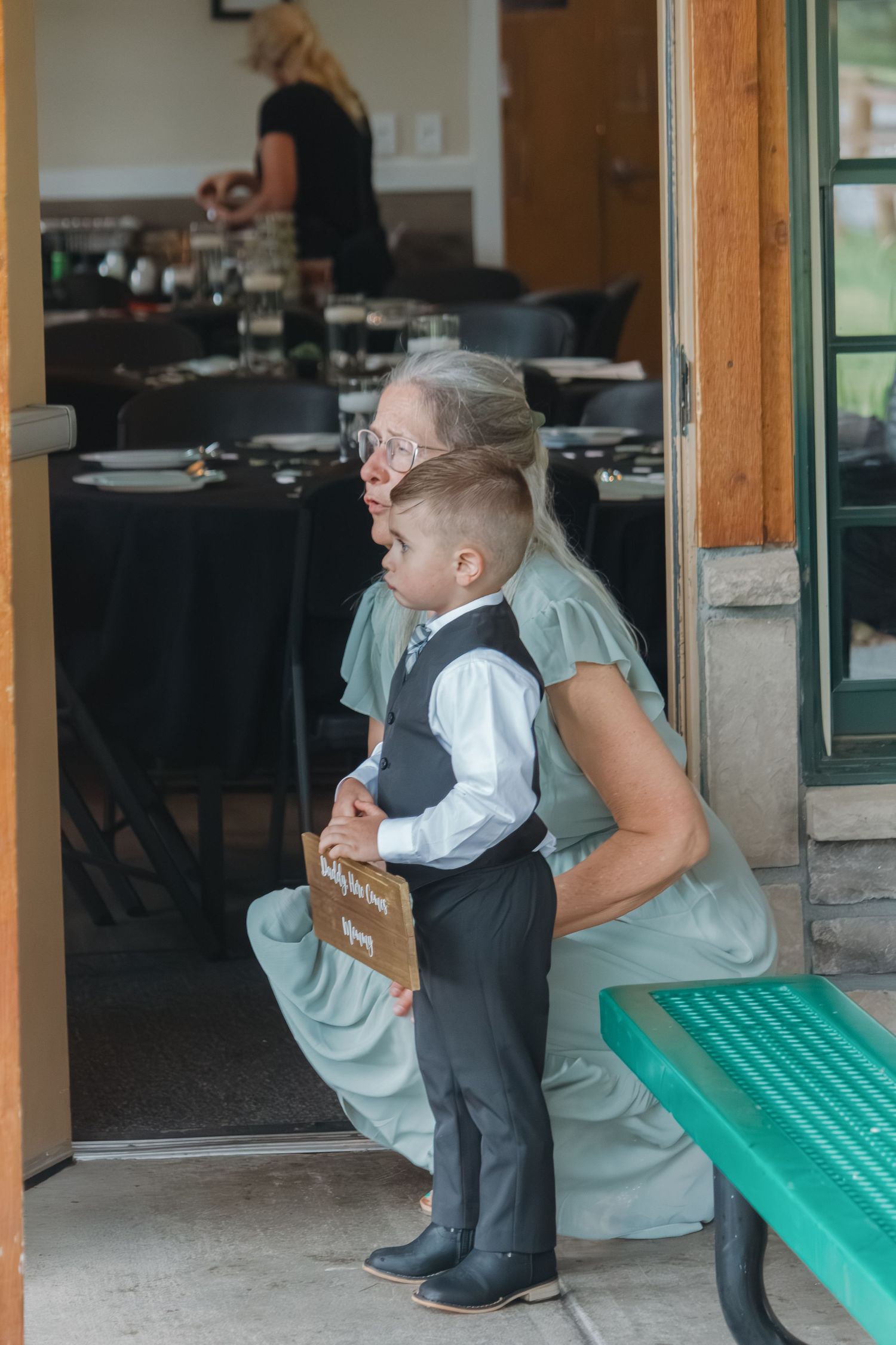 A small child in formal attire stands with support on a green bench outside a restaurant dining area.