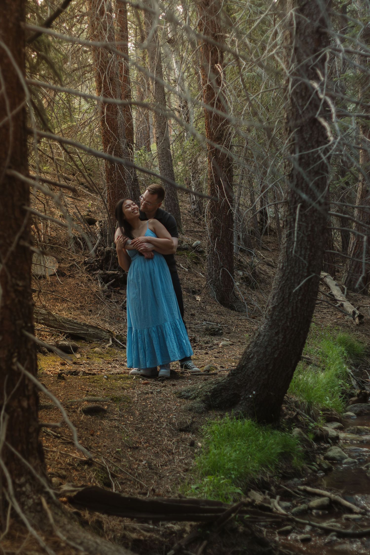 A loving couple shares a tender moment on a wooded trail surrounded by pine trees.