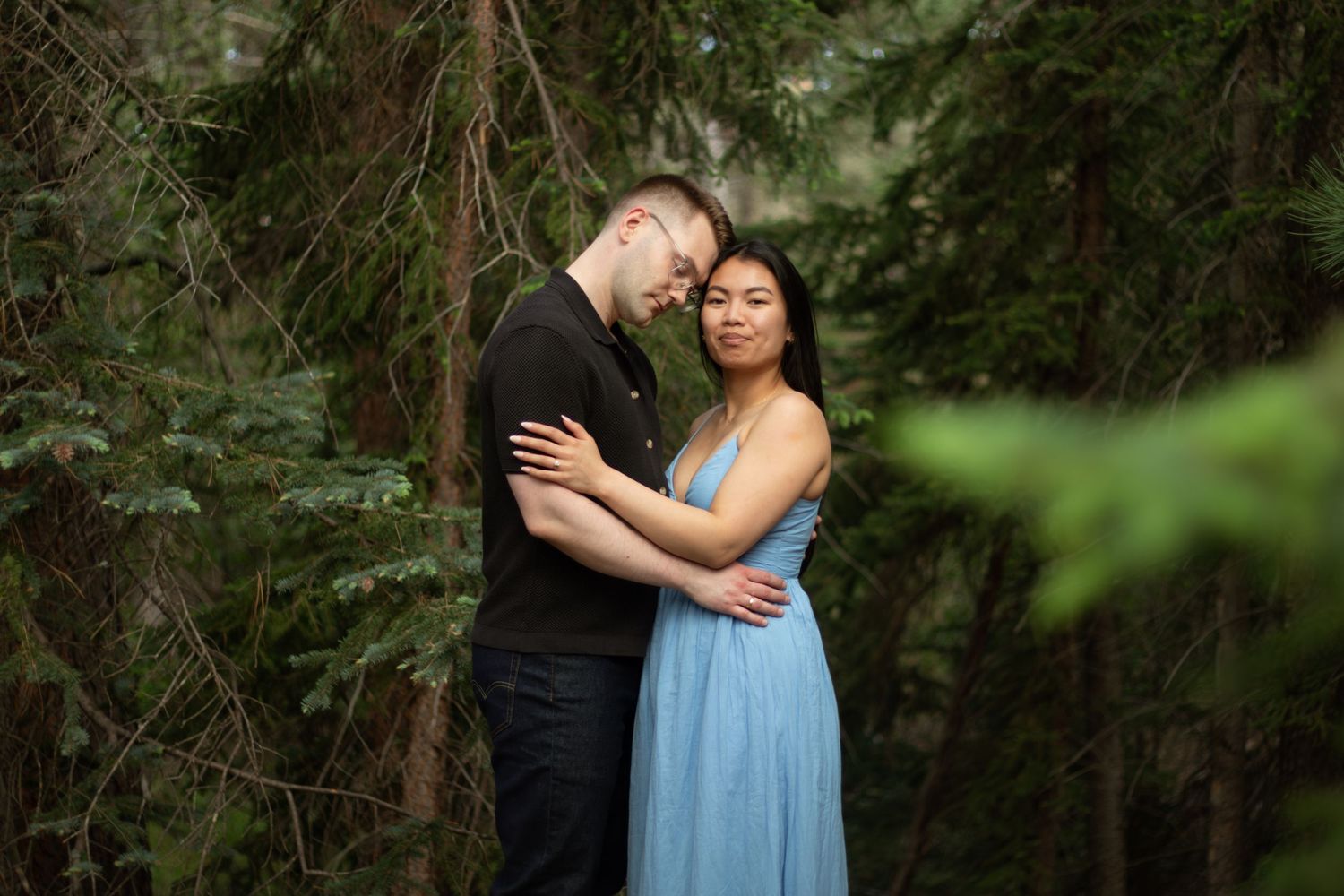 A couple holds each other close in a forest setting with soft natural lighting filtering through the trees.