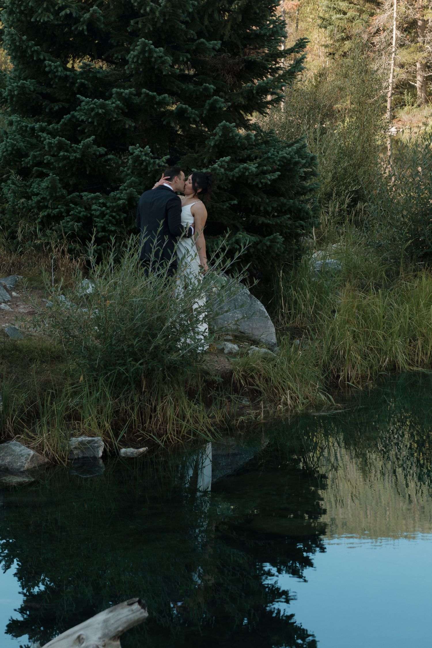 Newlyweds share a quiet moment by a reflective mountain lake at sunset.