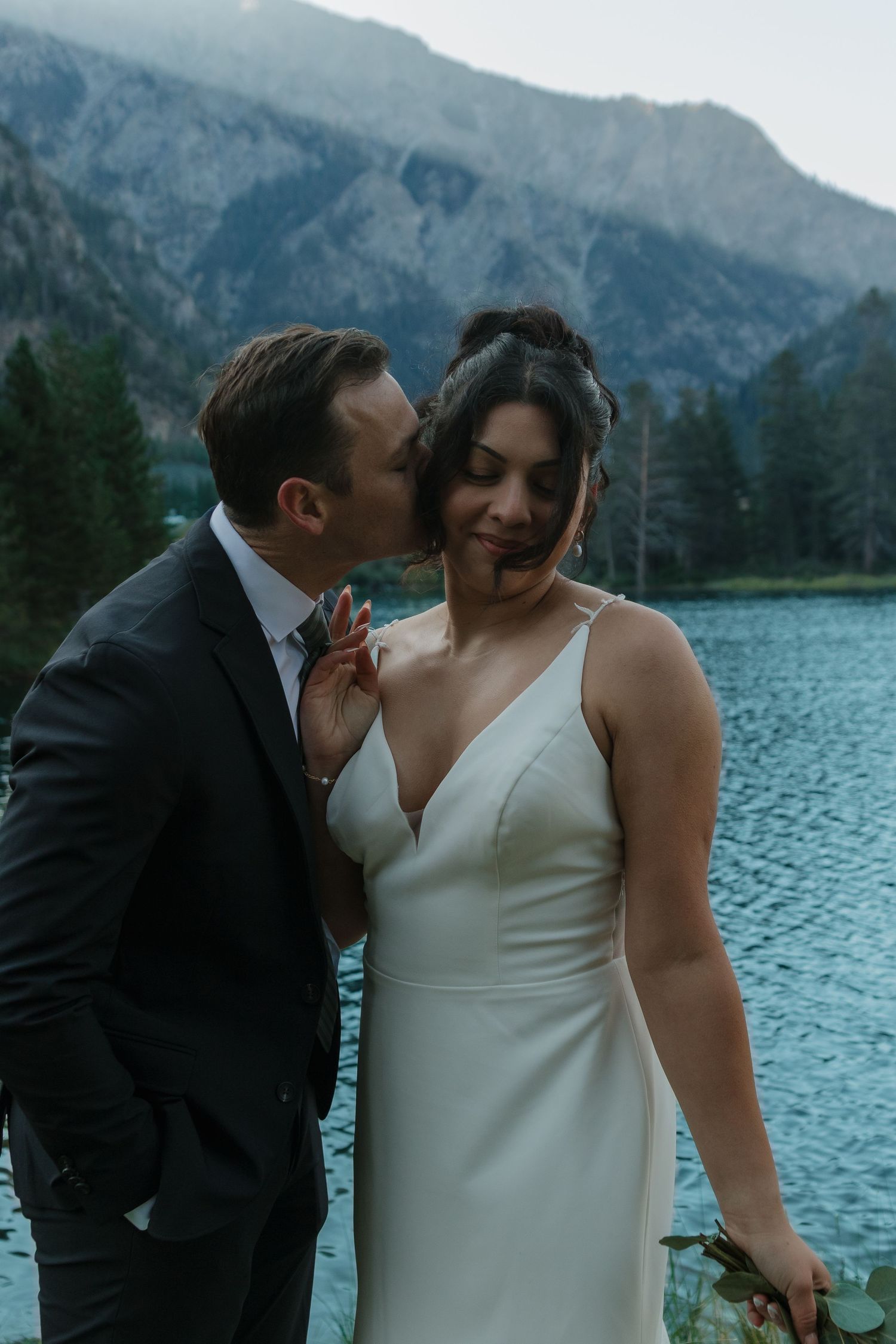 Intimate wedding moment captured by a scenic mountain lake with evergreen trees in background.