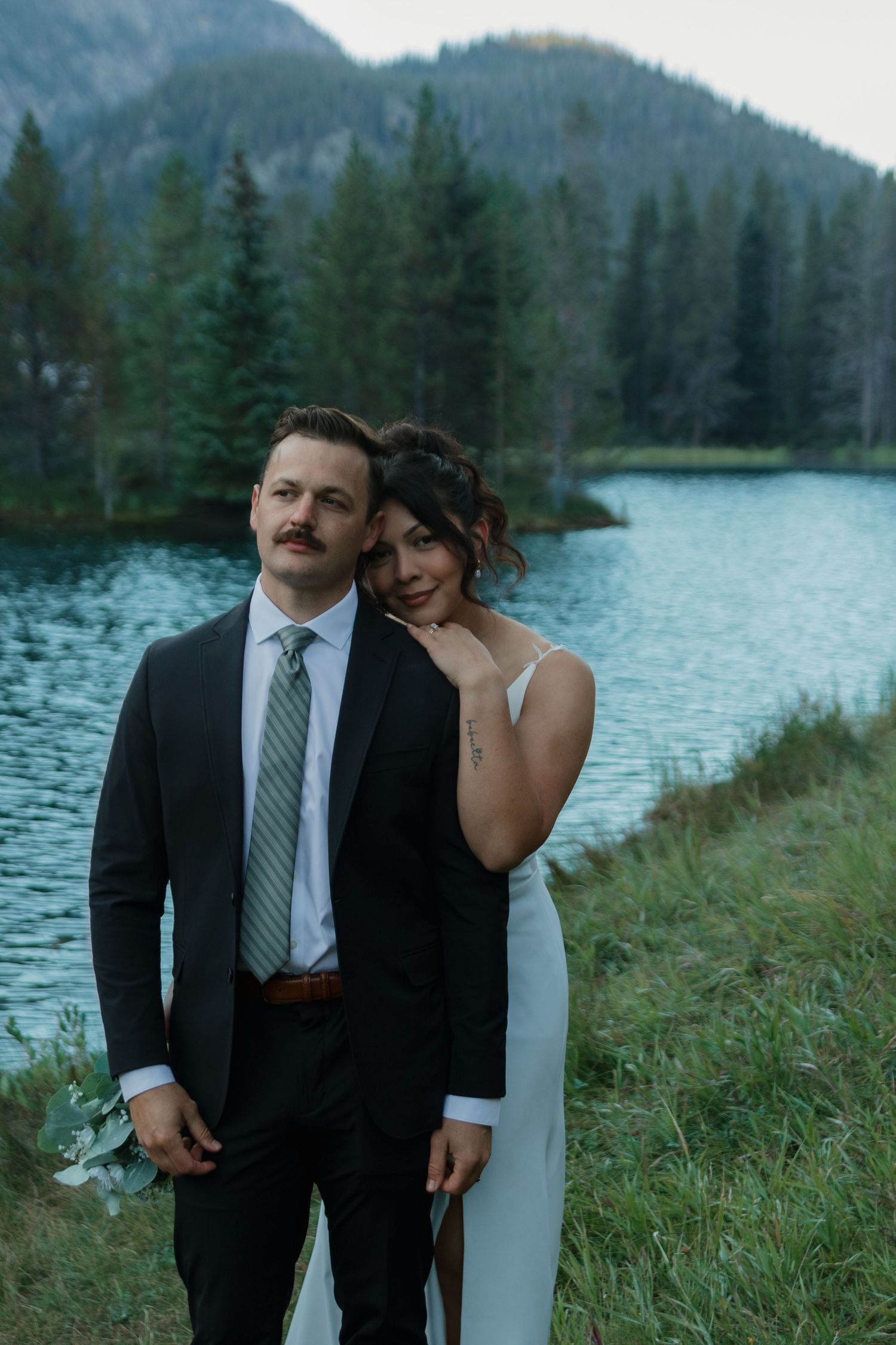 A romantic lakeside wedding photo with mountains and evergreen forest in the background.