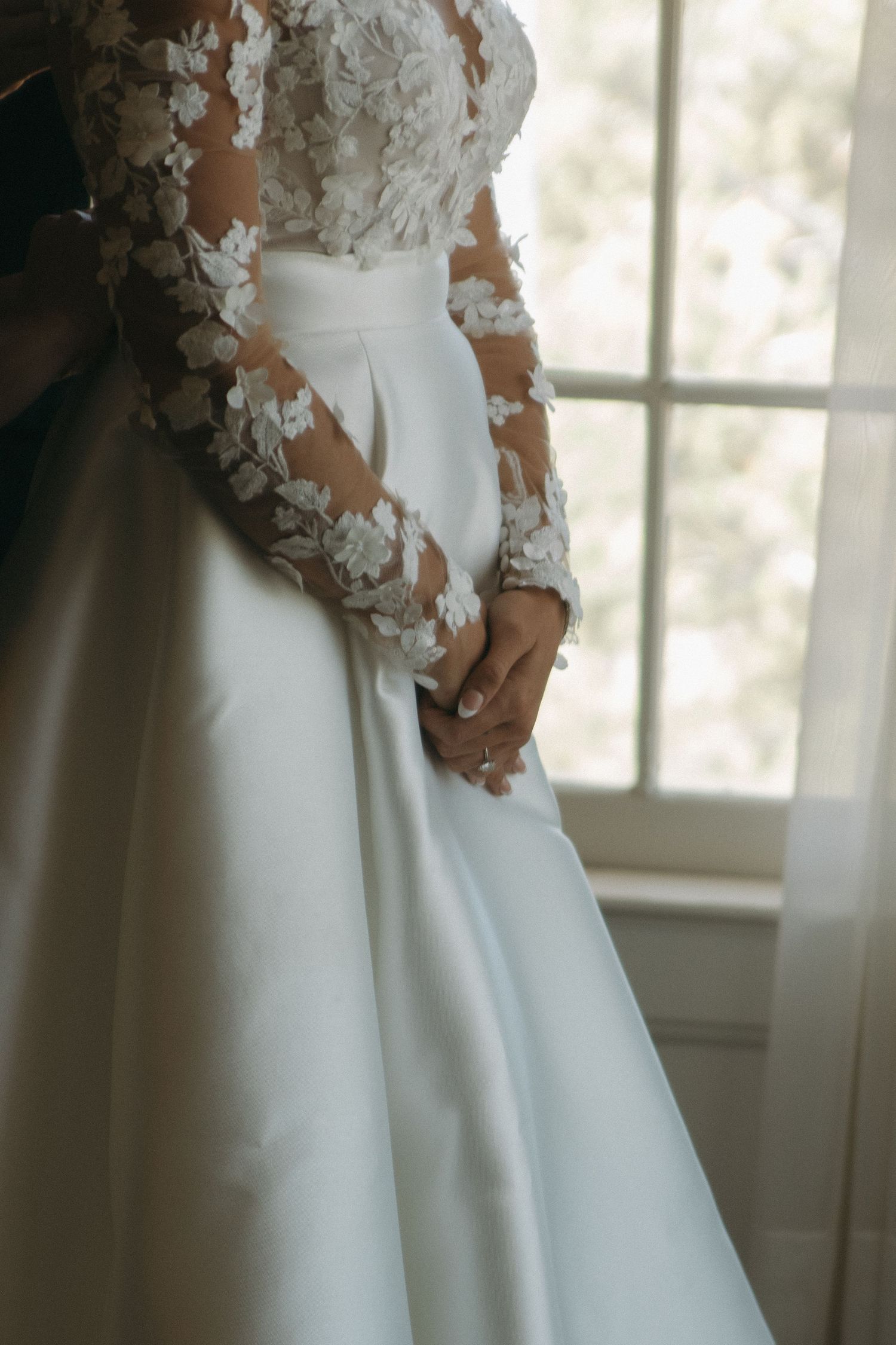 Close up detail of an elegant white wedding dress with delicate lace sleeves in natural window light.