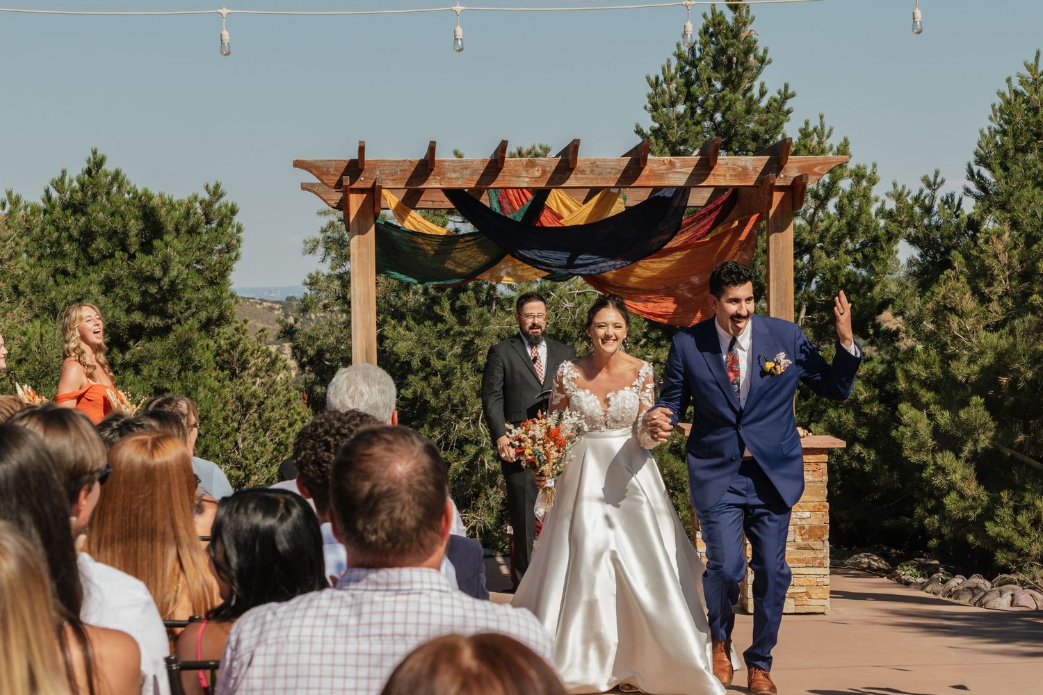 Outdoor wedding ceremony under a wooden pergola with guests seated on white chairs.