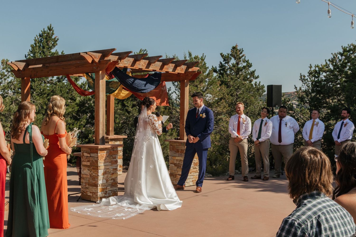 An outdoor wedding ceremony under a wooden pergola with guests watching the couple exchange vows.