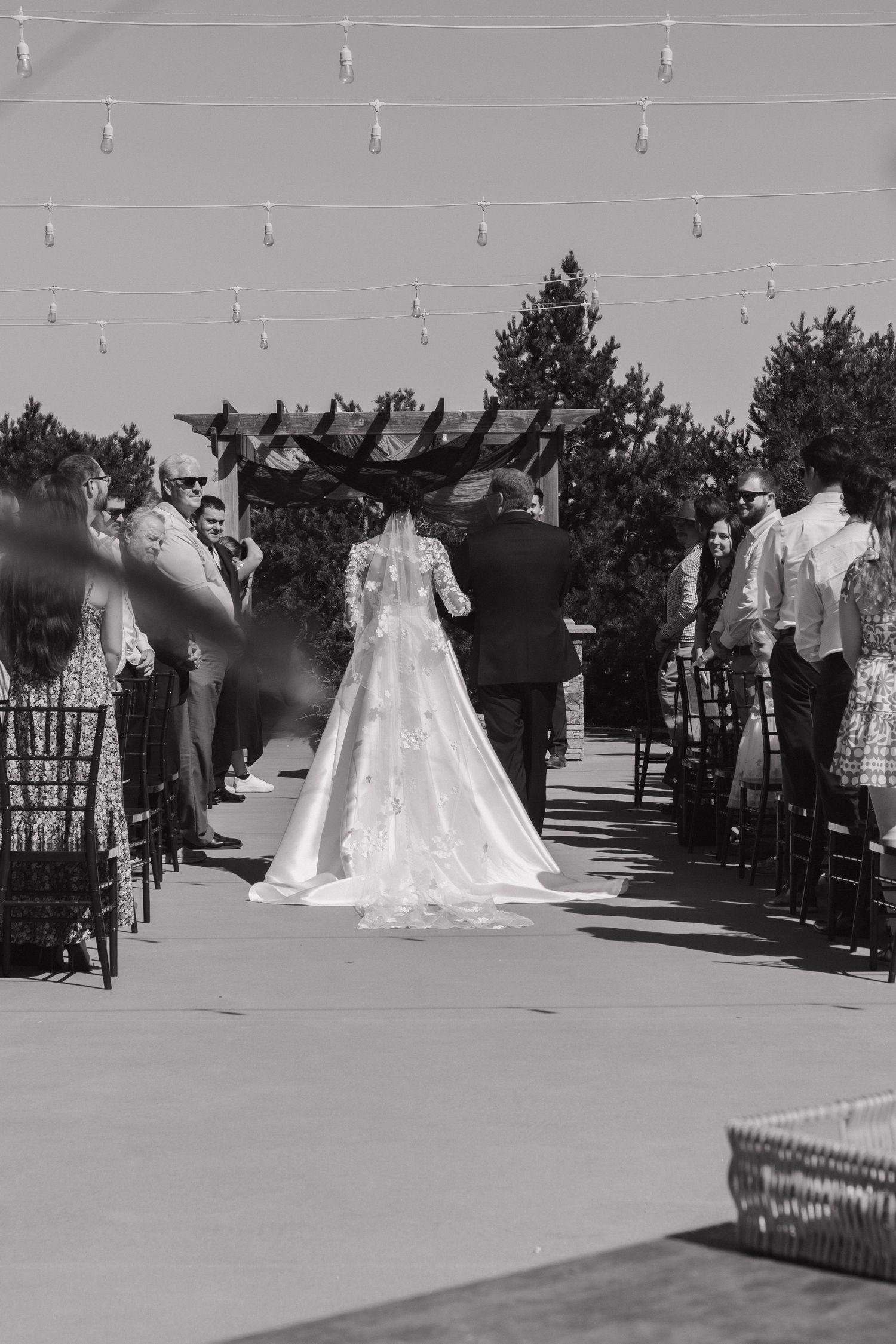Scenic outdoor wedding ceremony with white chairs and string lights overhead.