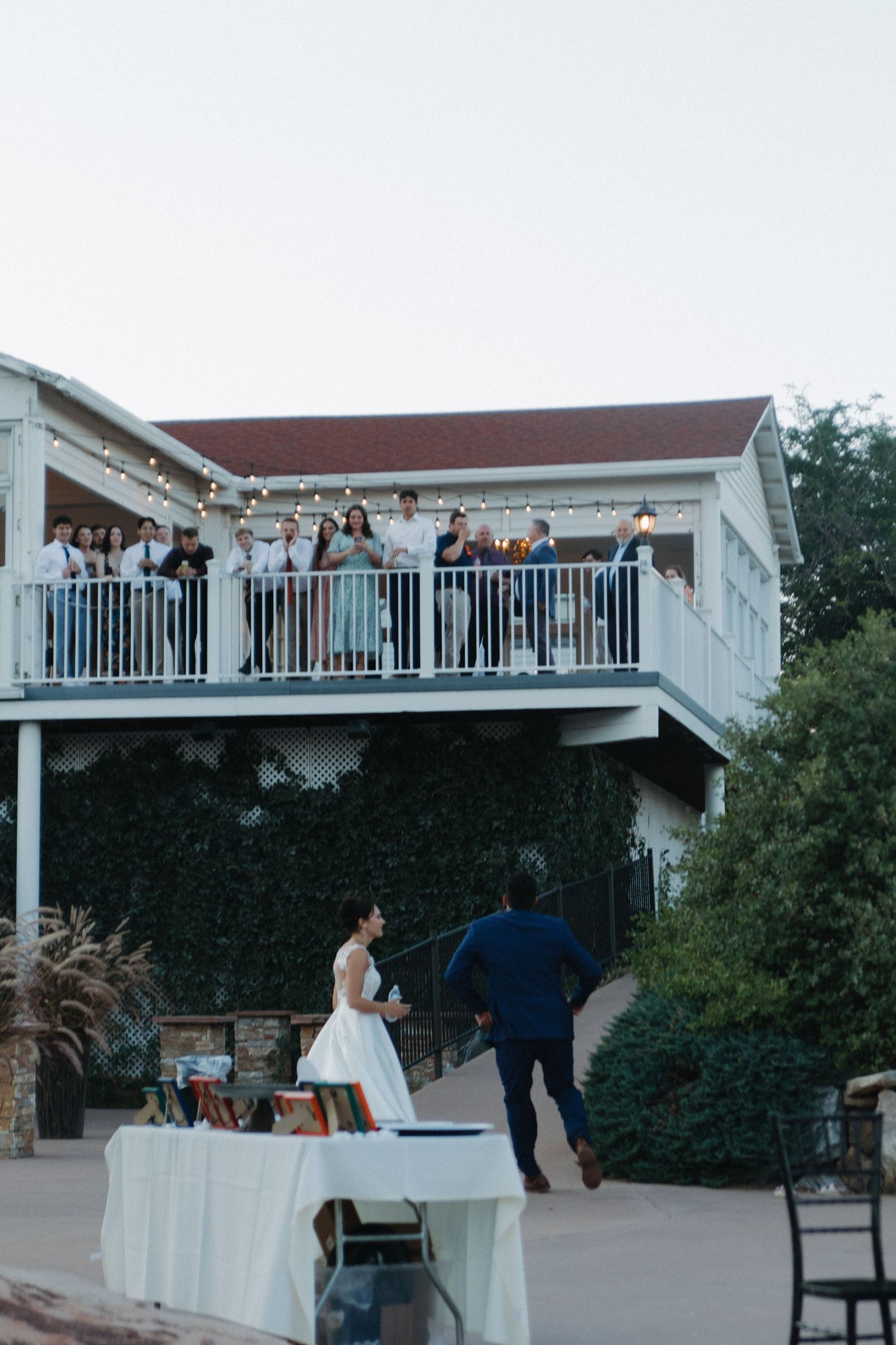 Wedding guests watch from a white balcony railing as festivities unfold on an outdoor terrace below.