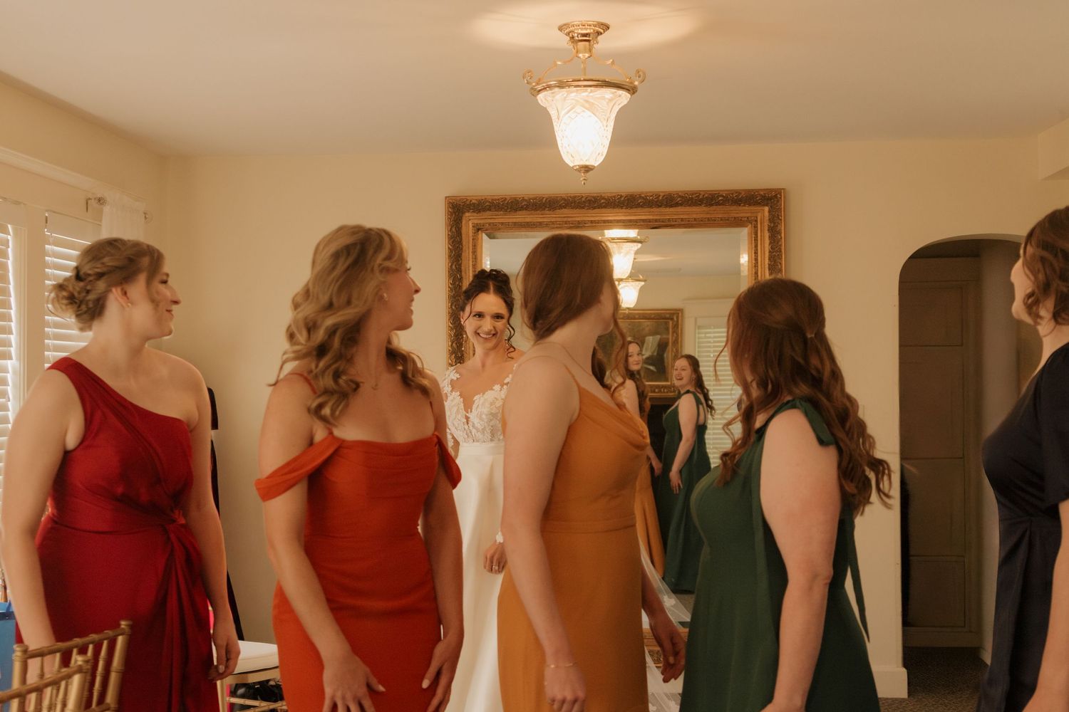 A group of women in colorful formal dresses gathered in a room with a gold-framed mirror and warm lighting.