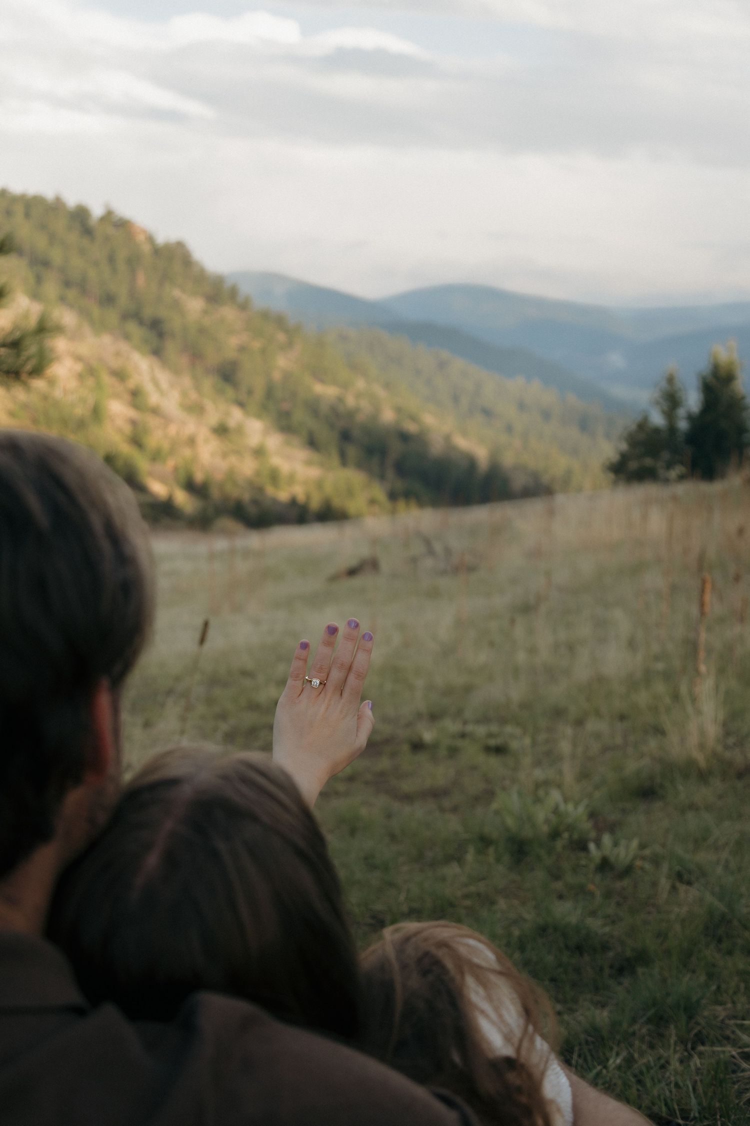 A woman shows off her engagement ring with a scenic mountain valley view behind her.