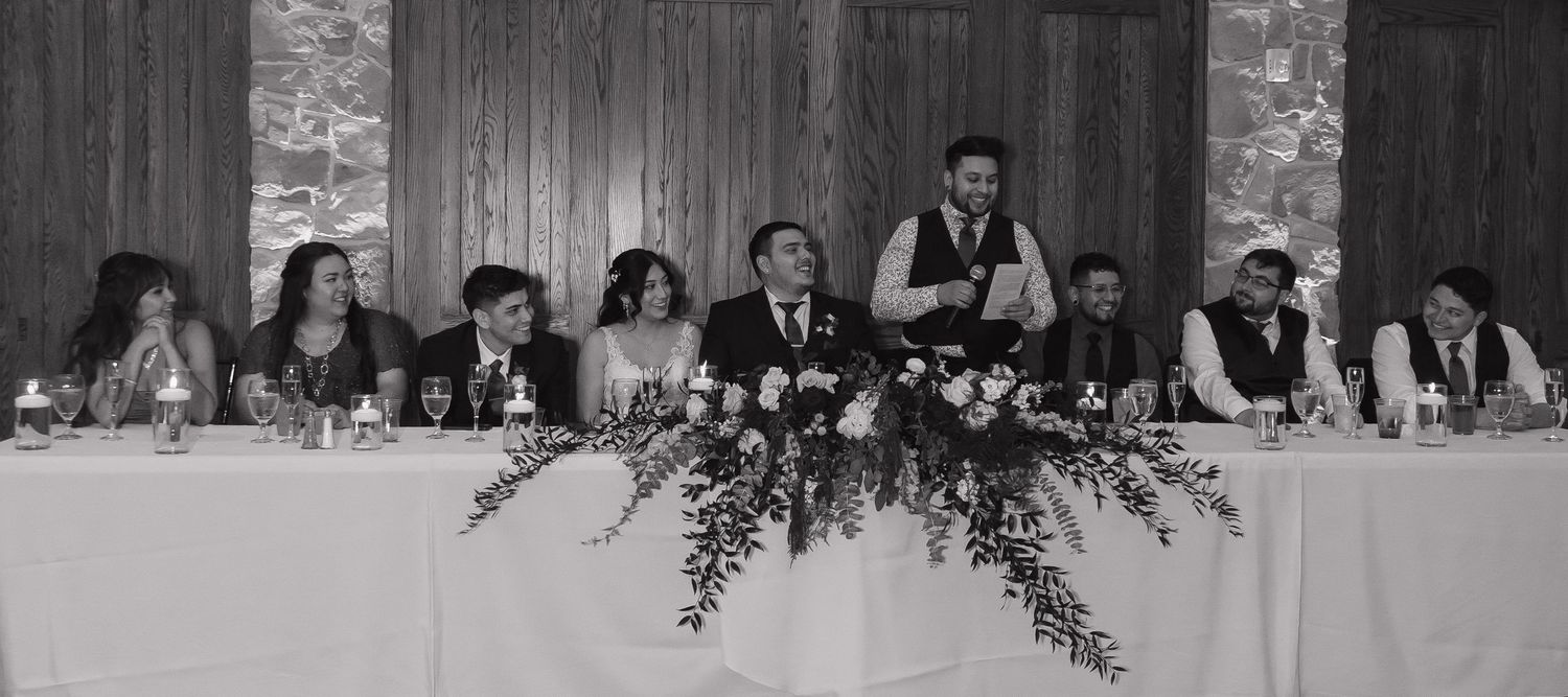 Wedding party seated at head table with floral centerpiece during reception in black and white photo.
