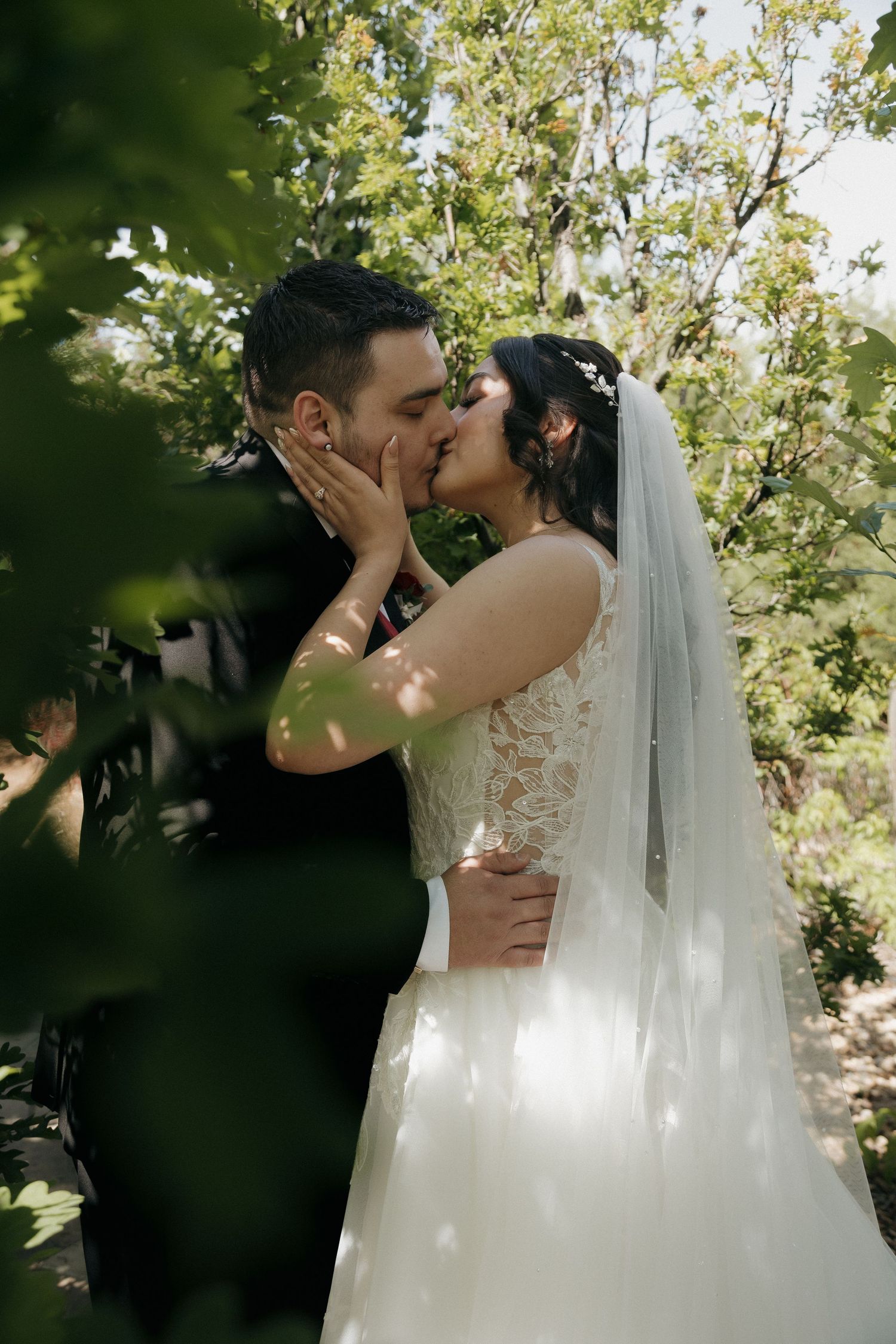 Bride and groom sharing a romantic kiss surrounded by green foliage on their wedding day.
