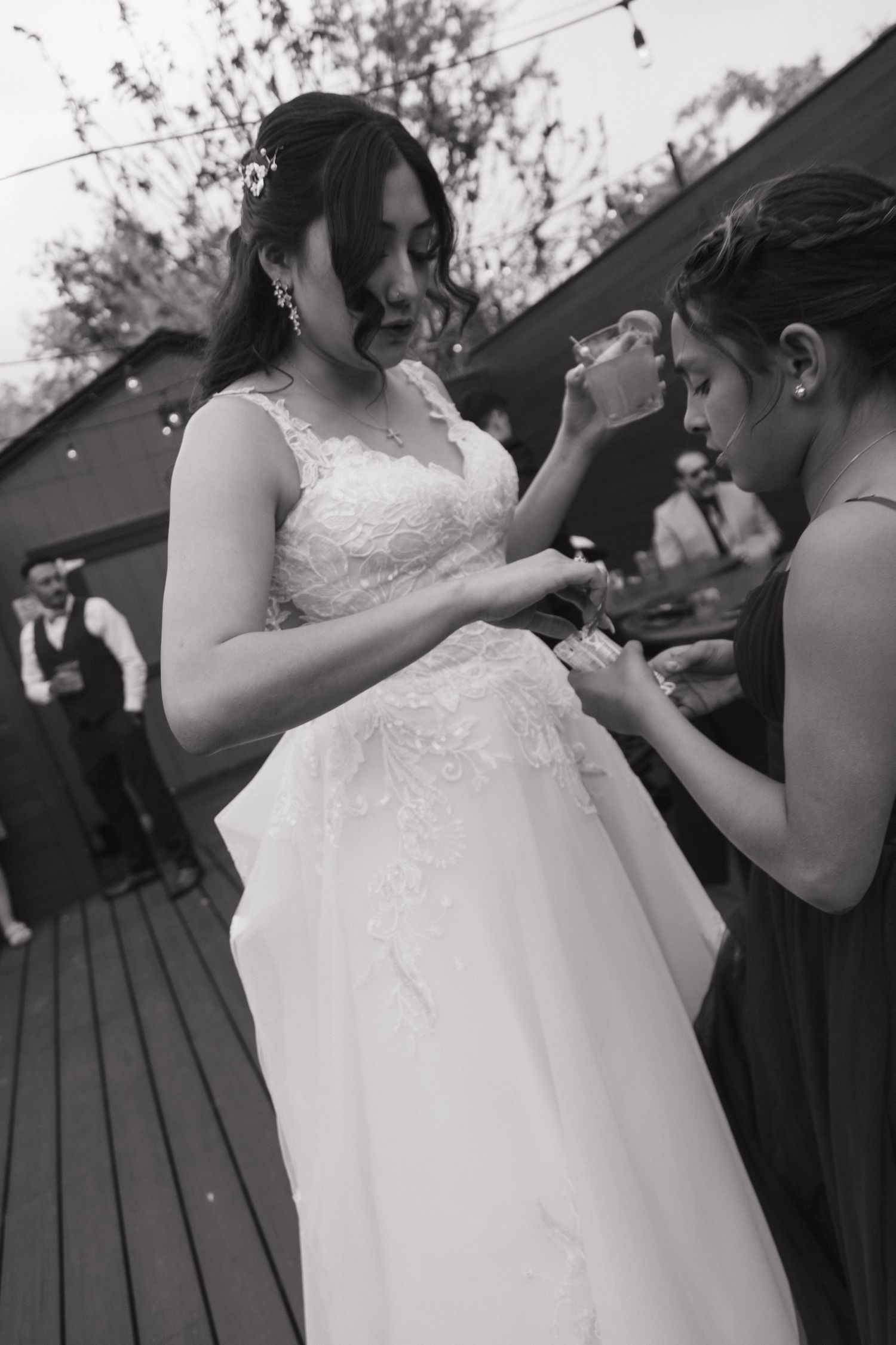Black and white photo of bridesmaid helping bride with her dress at outdoor wedding reception.