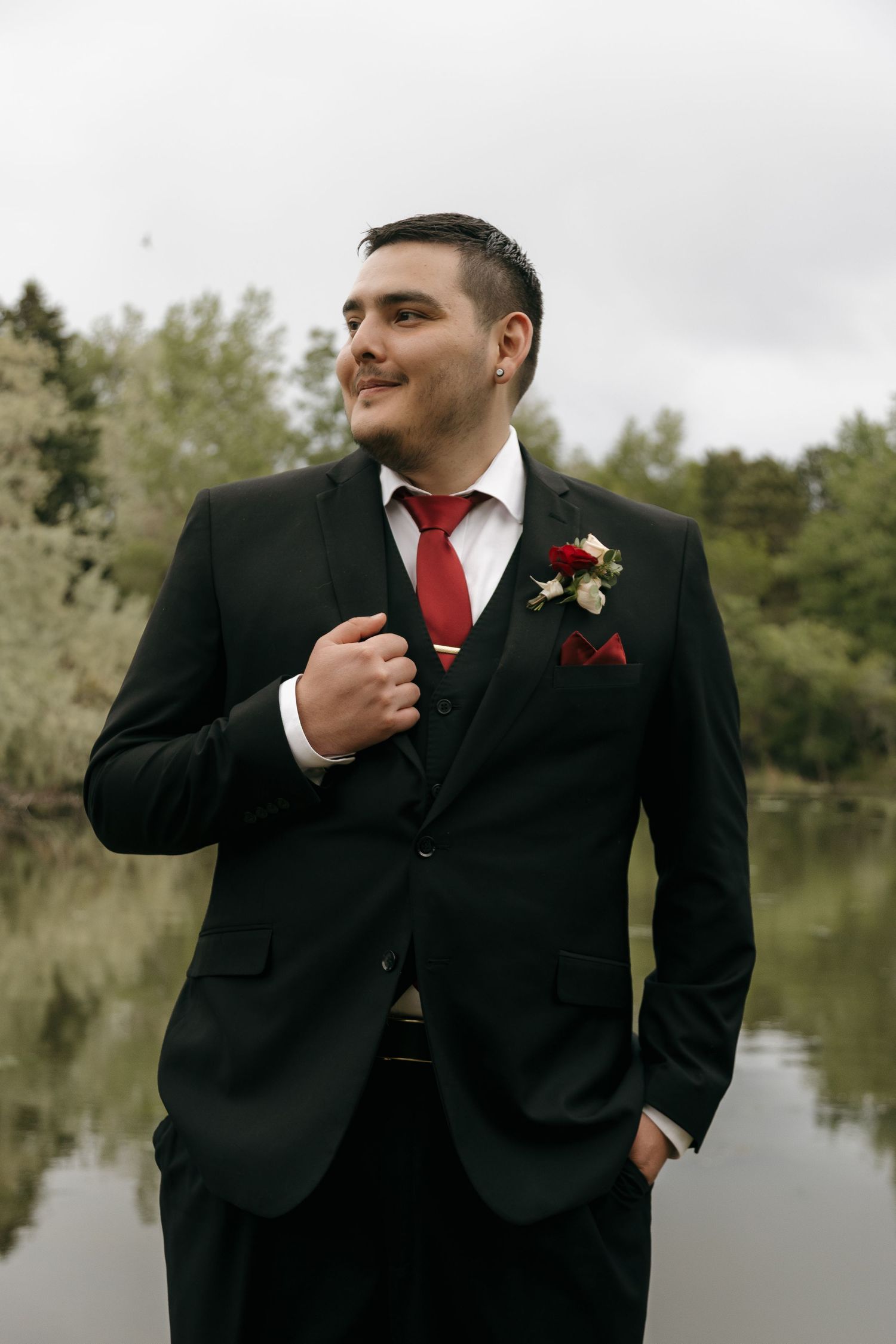 Handsome groom in black suit with red tie and floral boutonniere smiling confidently outdoors near a serene lake.