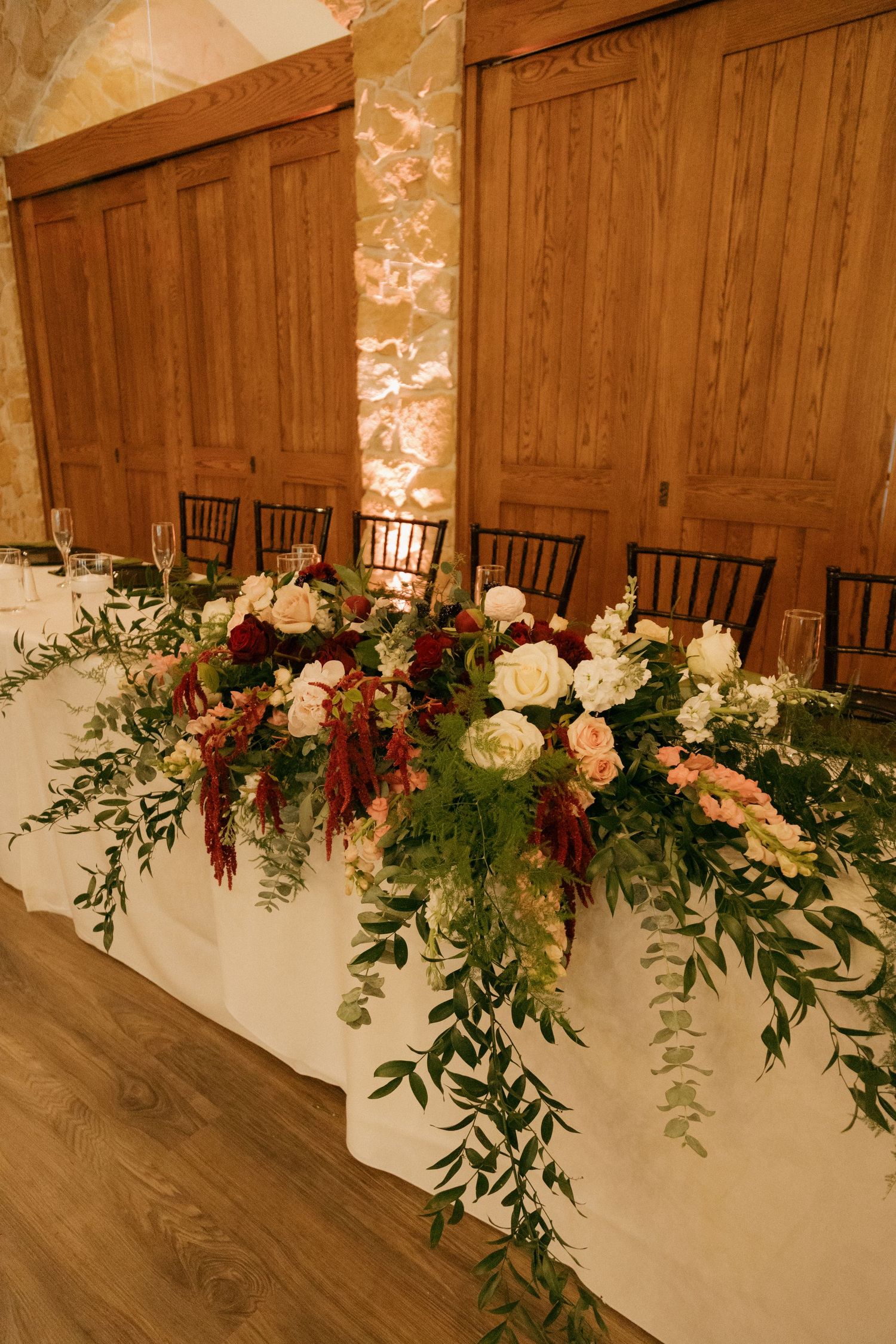 Elegant wedding head table adorned with lush floral arrangement featuring burgundy, blush, and white roses with cascading greenery.