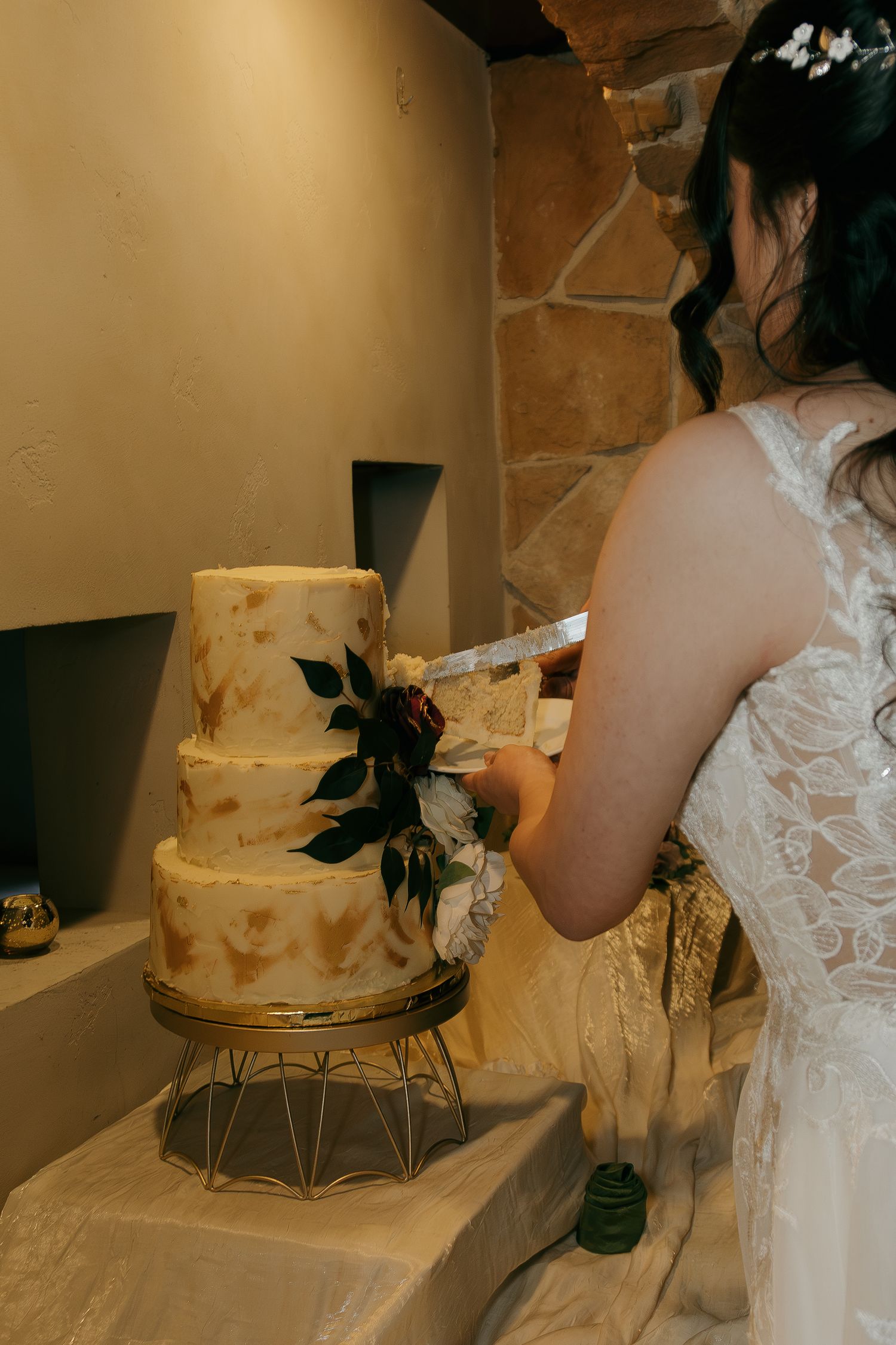 Bride cutting three-tier white wedding cake decorated with flowers at reception.