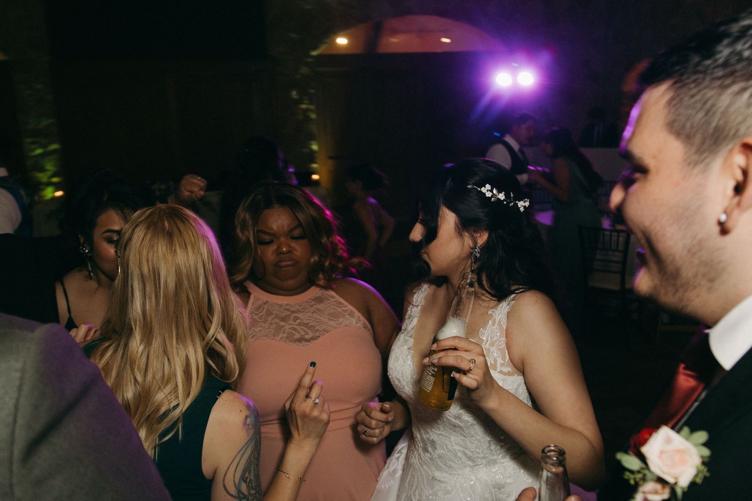 Bride in white lace gown dancing with guests under purple lights at lively wedding reception.