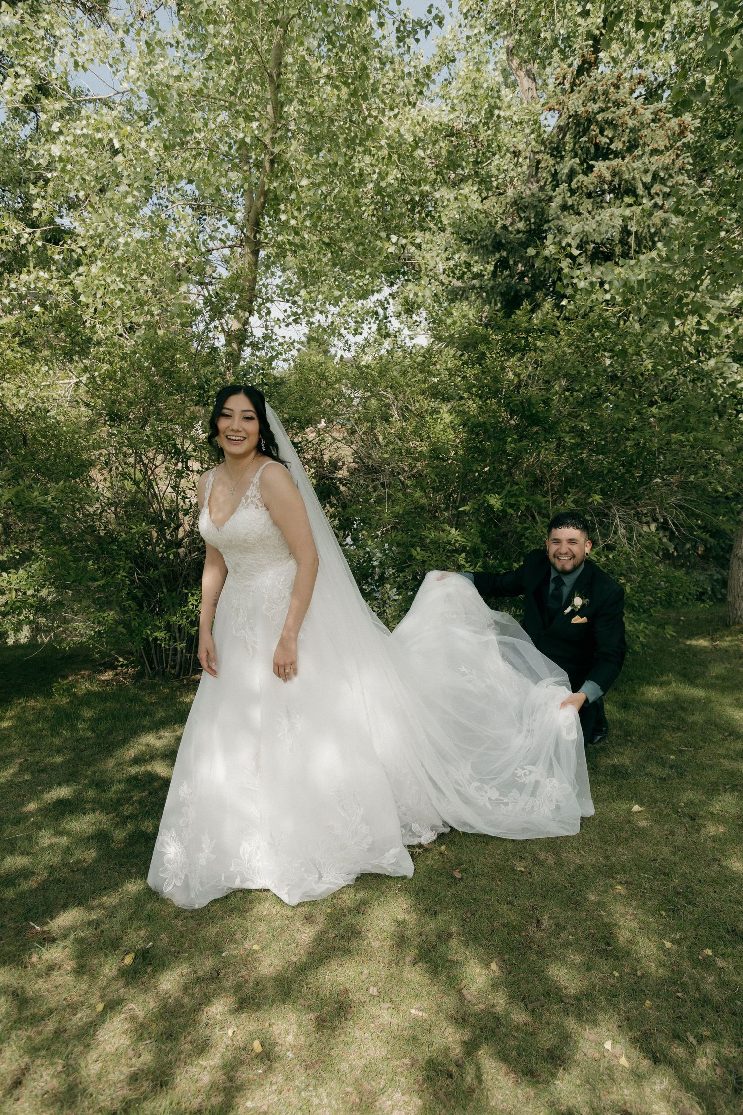 A smiling bride in a white gown and veil poses with the groom in a lush green garden.