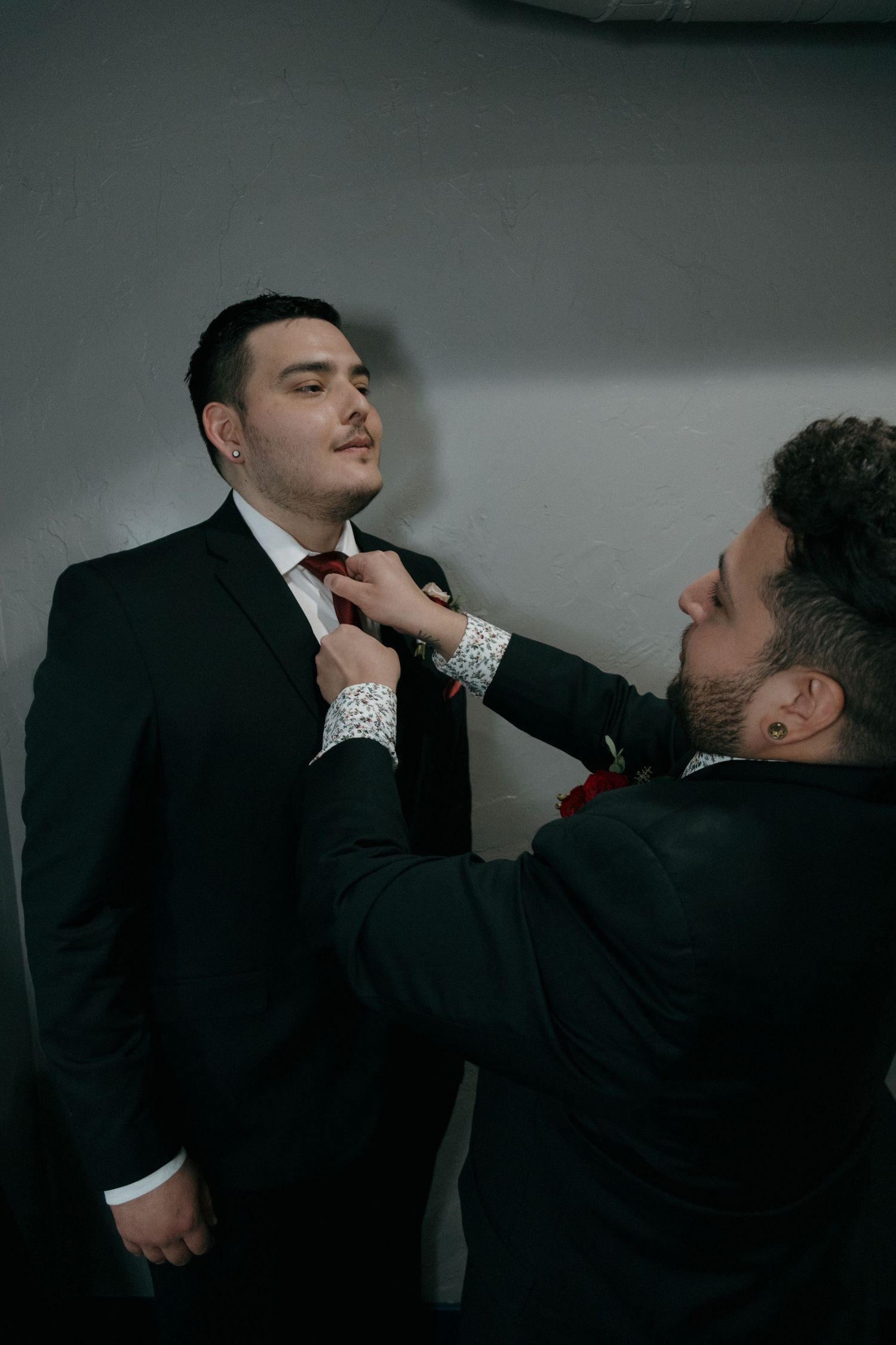Groomsman helping groom adjust his tie while both wear black suits before wedding ceremony.