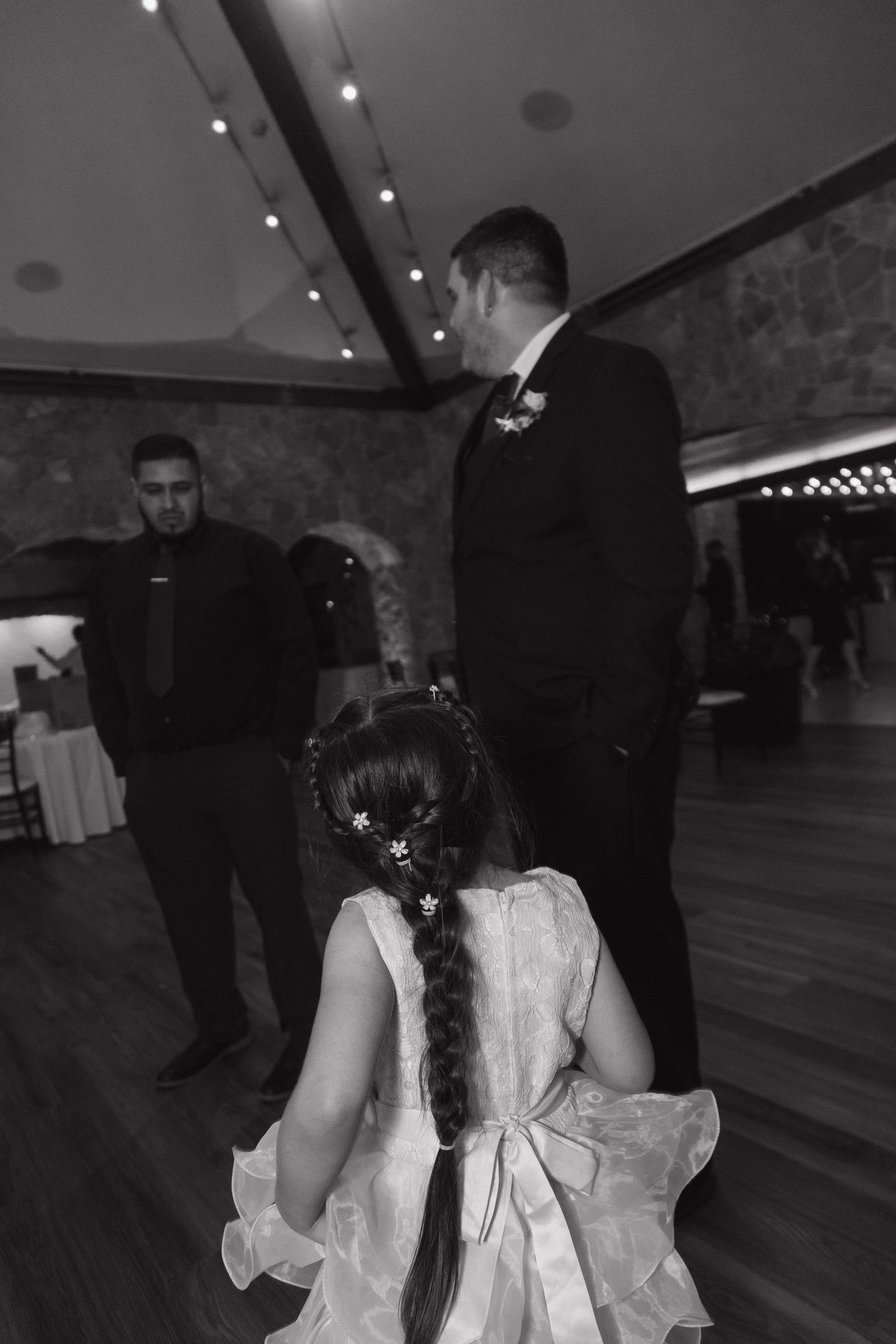 Black and white photo of flower girl with braided hair approaching groom on wedding dance floor.