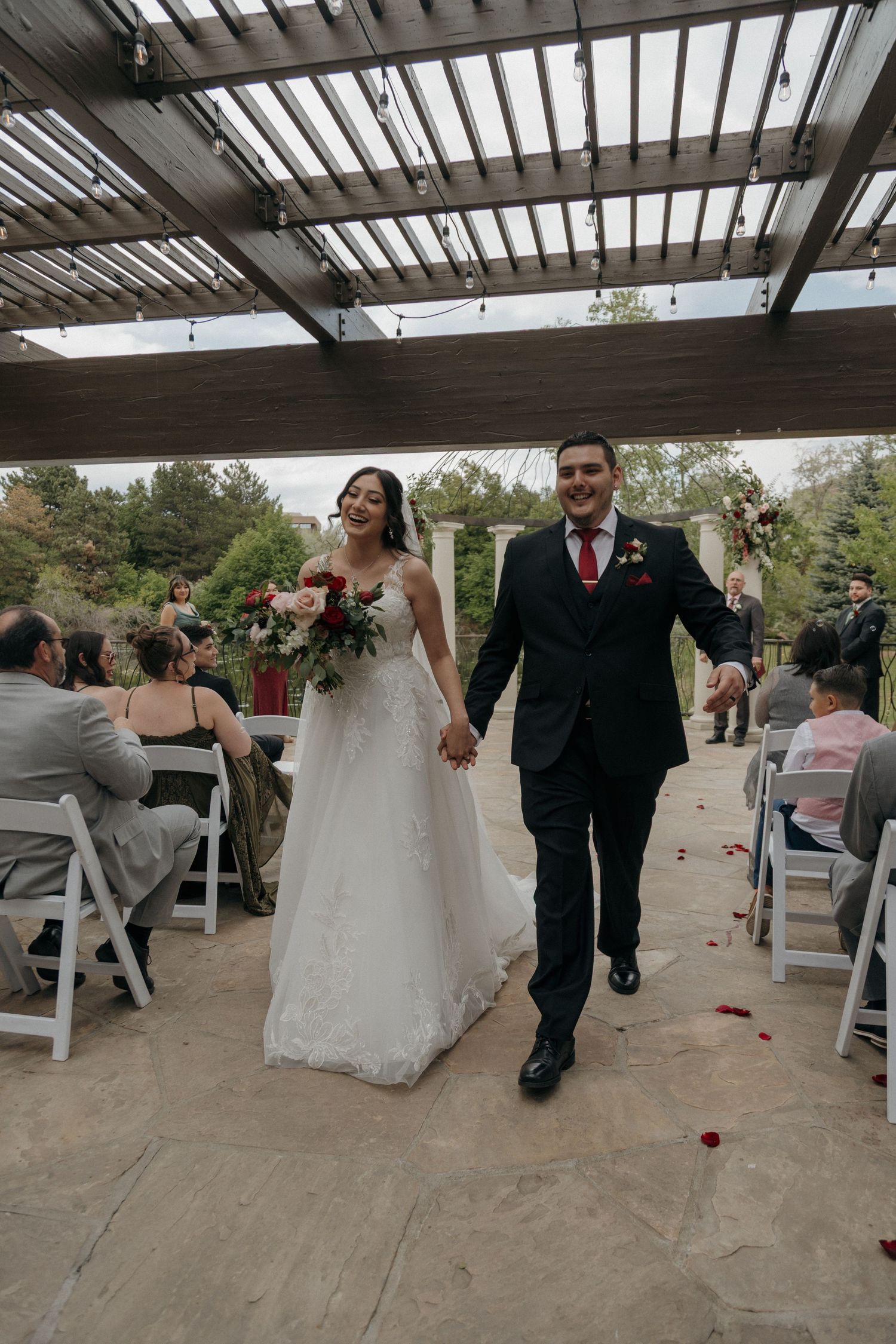 Newlyweds walk hand-in-hand down aisle after outdoor wedding ceremony under pergola with guests seated.