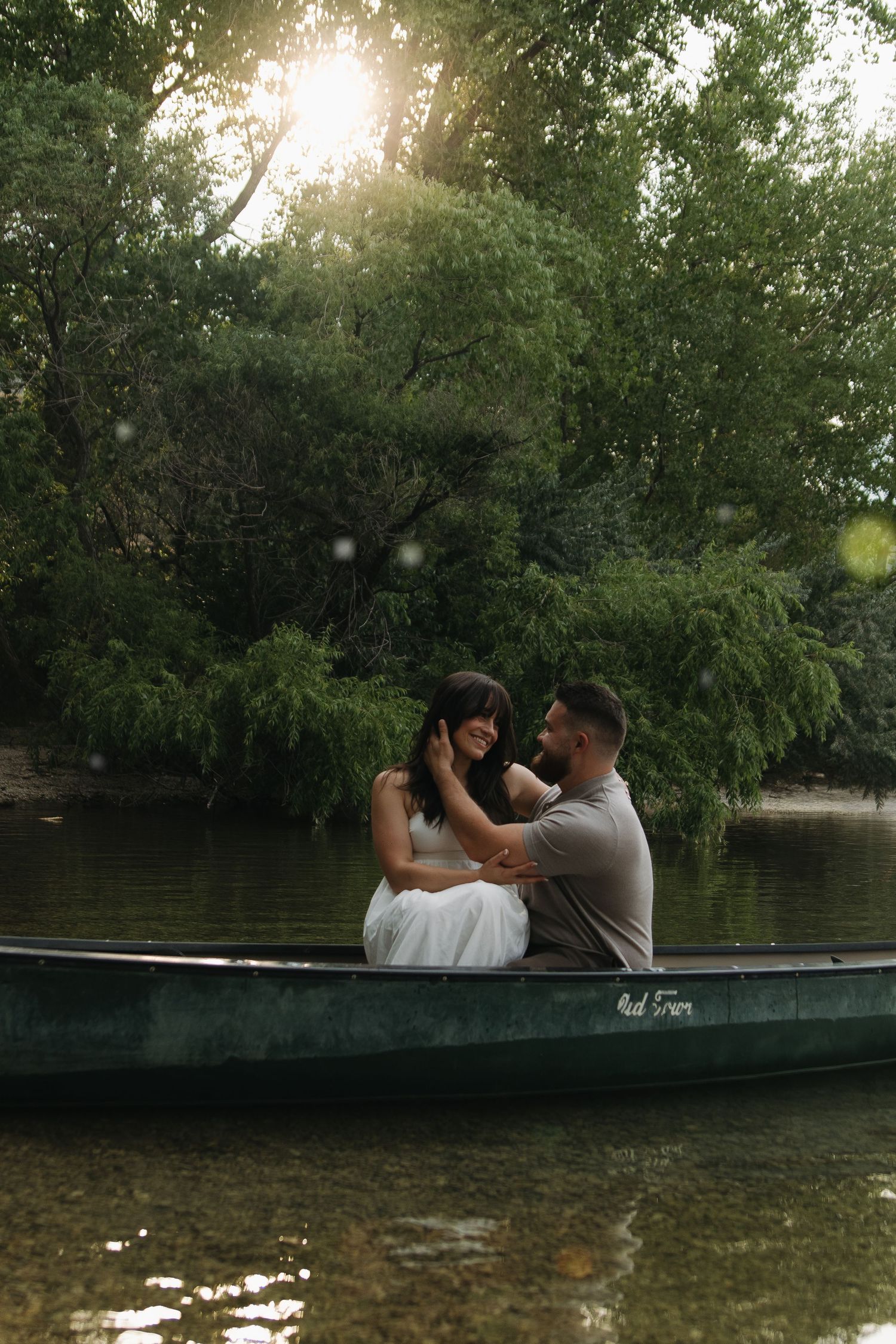 Couple sitting intimately in a green canoe on a calm river surrounded by lush trees with soft sunlight filtering through.