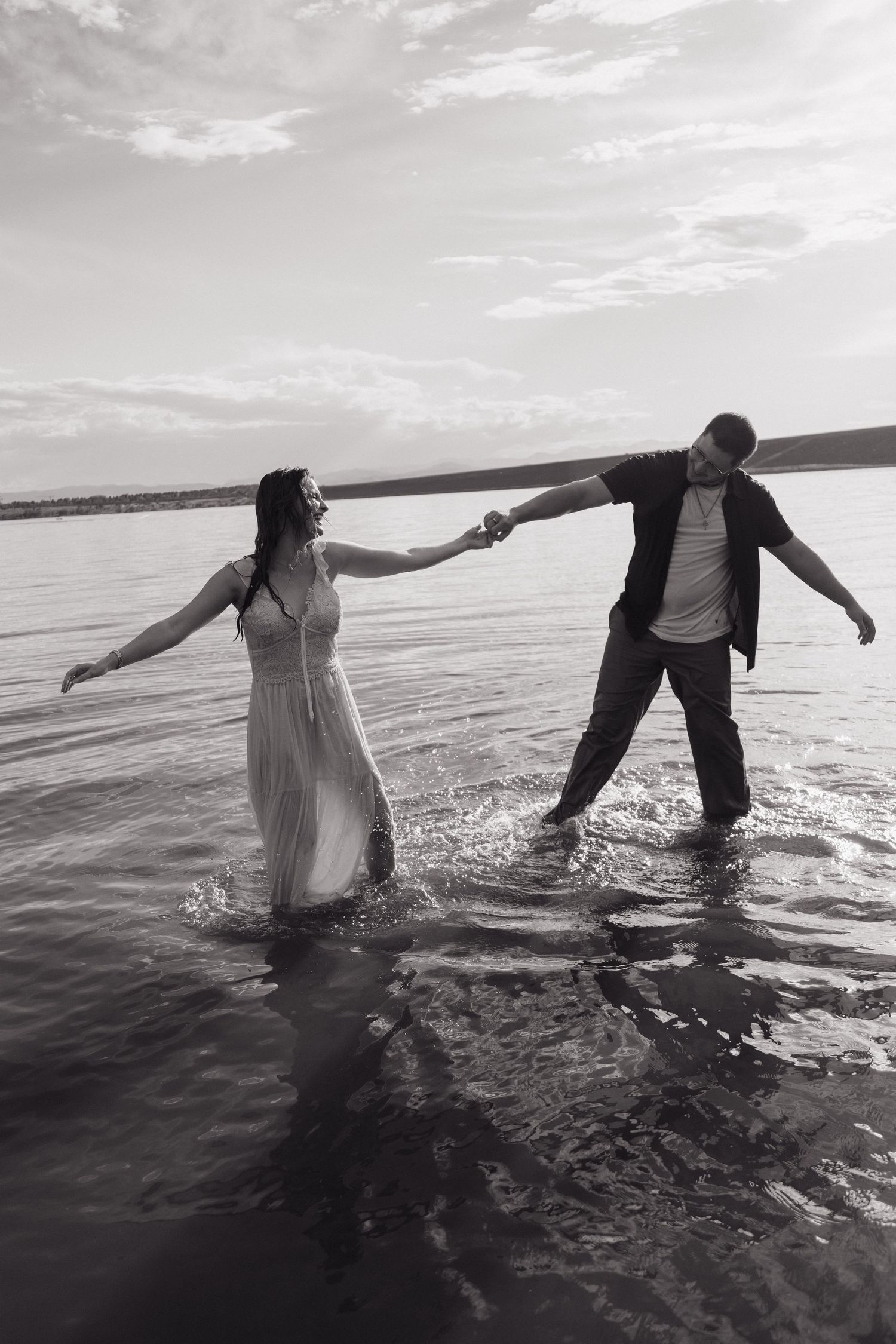 Black and white photo of couple holding hands while wading through lake water together.