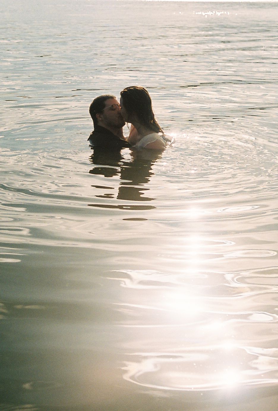 Couple sharing a romantic kiss while standing in sunlit shimmering water outdoors.