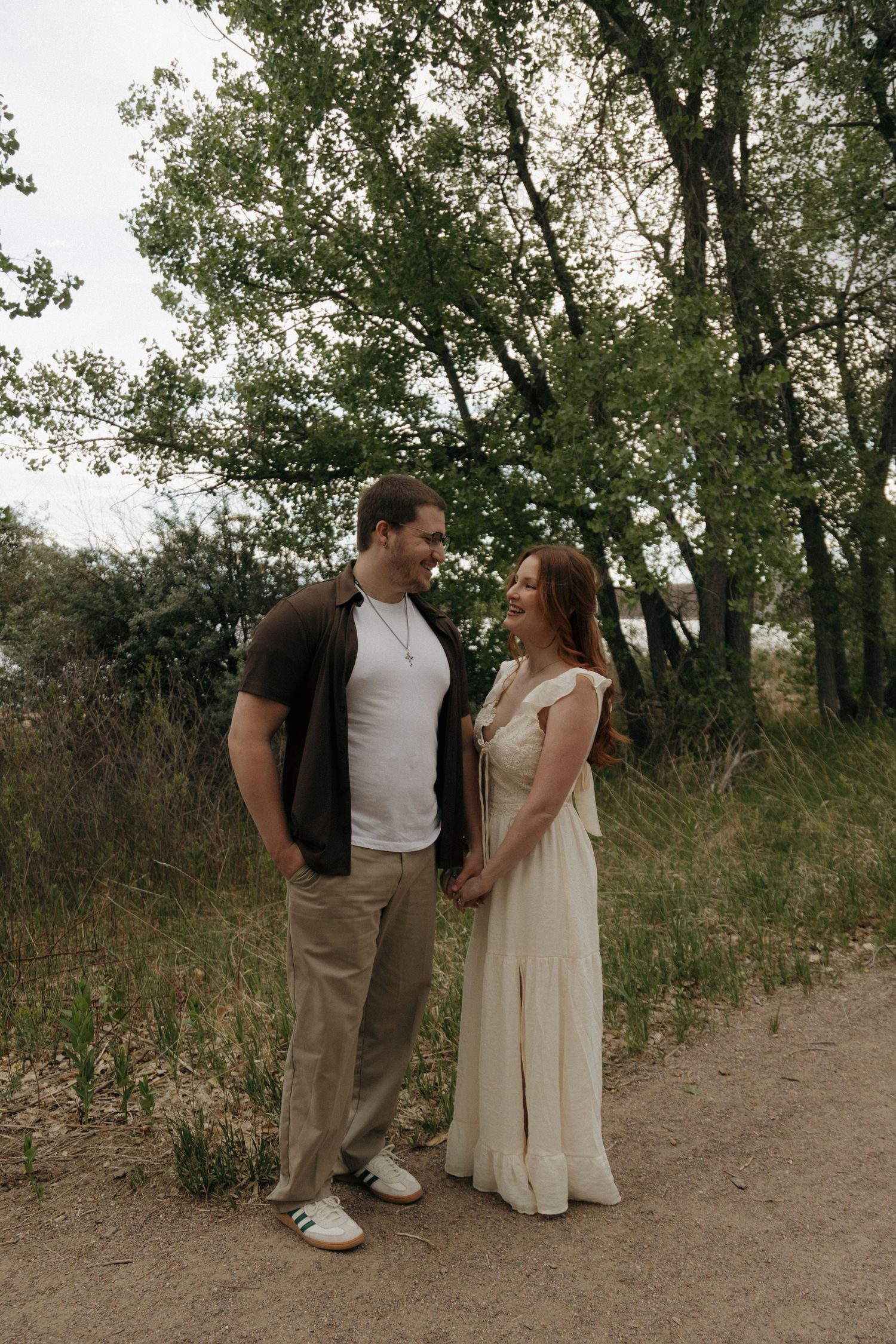 Couple holding hands and smiling at each other on a nature trail surrounded by lush green trees.