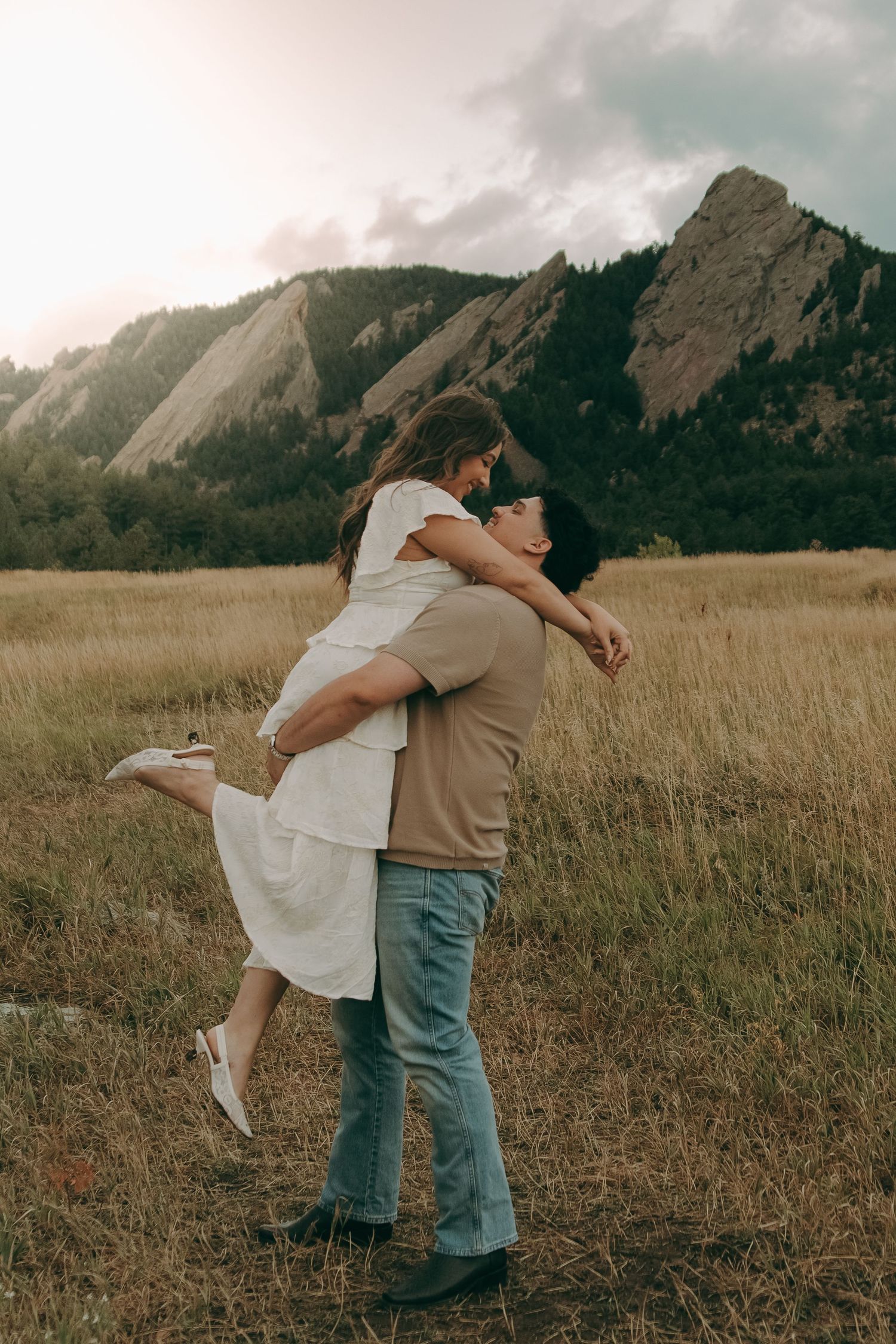 Groom lifts bride in white dress joyfully in sunlit meadow with stunning Colorado Flatirons behind them.