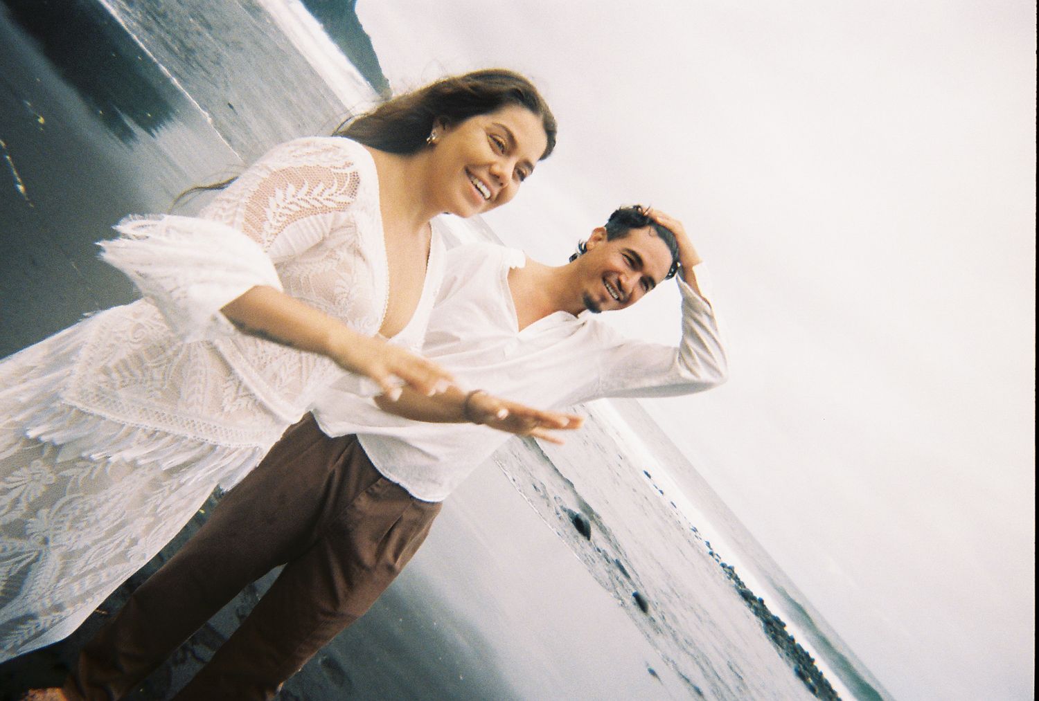 Joyful couple holding hands and leaning back playfully near ocean shoreline on film photo.