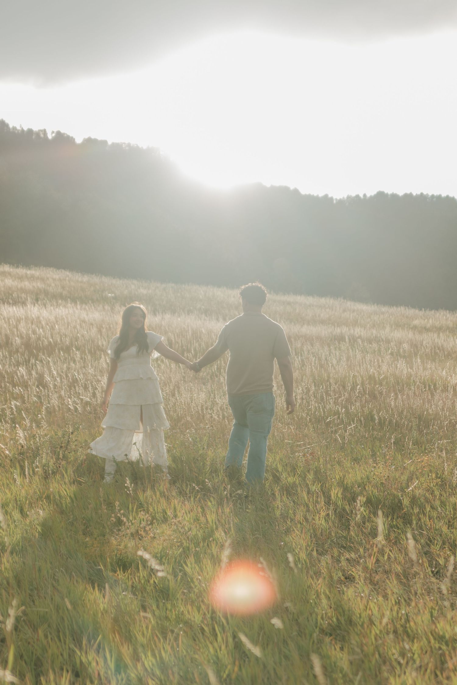 Couple holding hands walking through a golden field toward misty mountains in warm sunset light.
