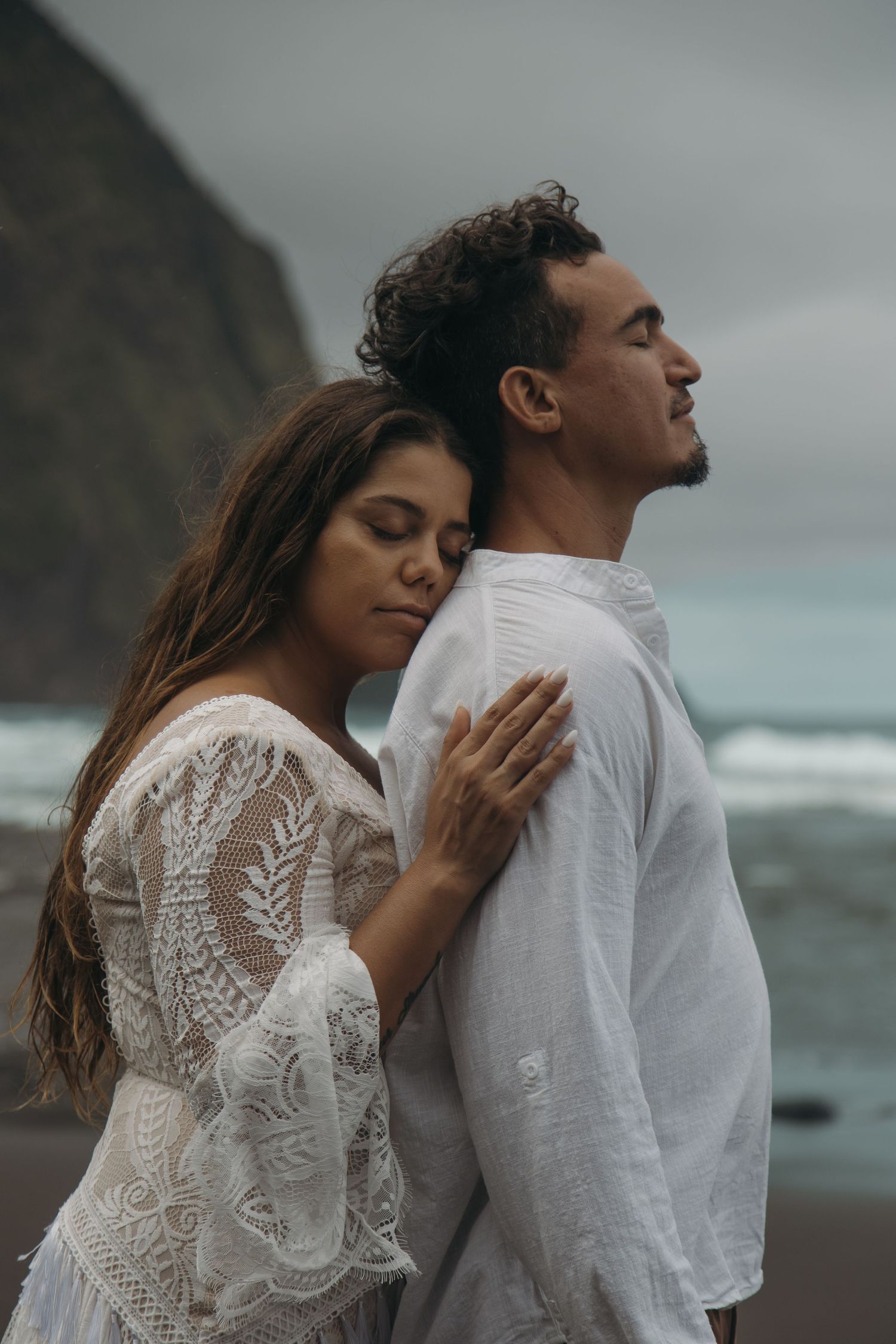 Couple in white outfits embracing on a black sand beach with dramatic cliffs and ocean waves in the background.