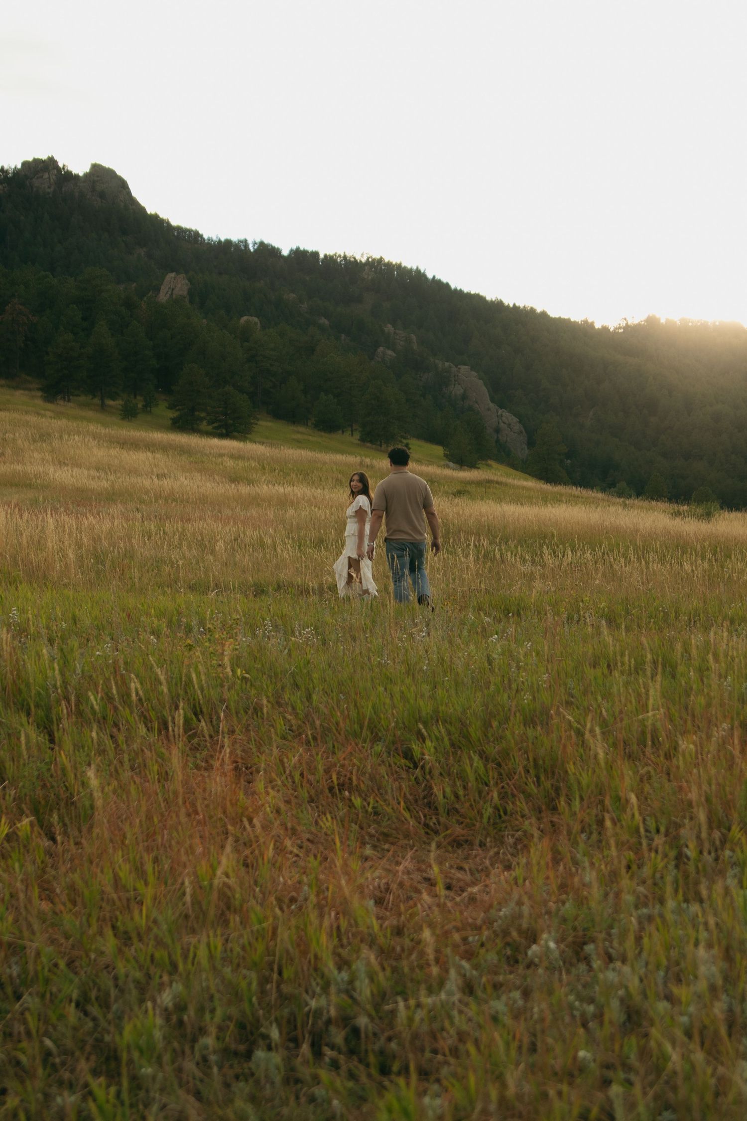 Couple walking hand in hand through a lush green meadow toward forested hills in golden sunset light.