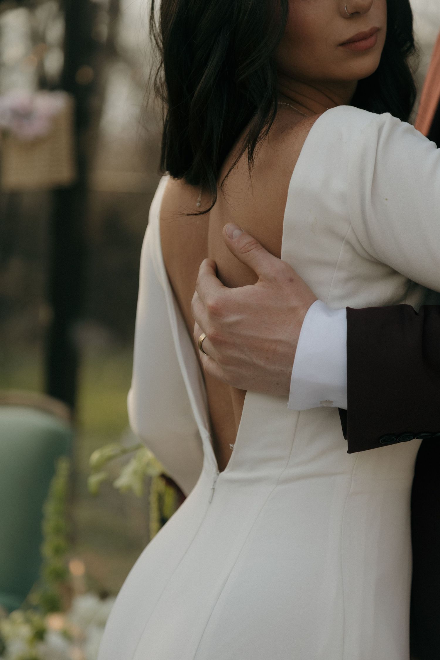 Groom in black suit embracing bride in elegant open-back white wedding gown outdoors.