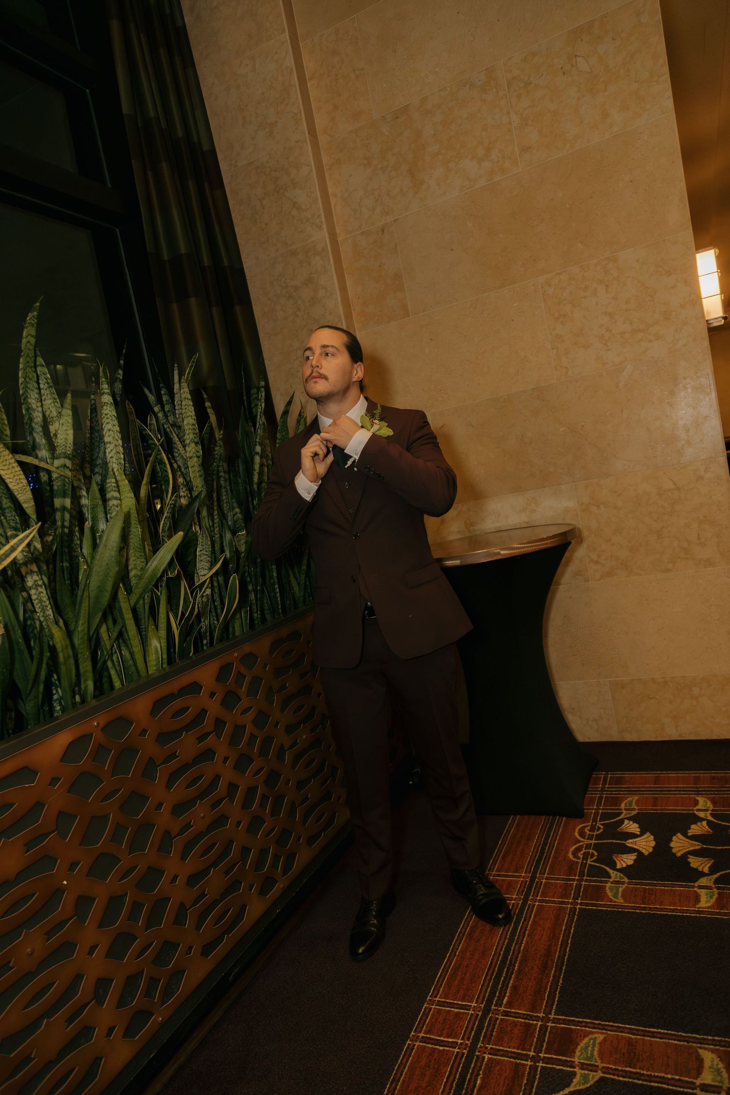 Groom in brown suit adjusting tie in elegant hotel lobby with green plants and warm lighting.