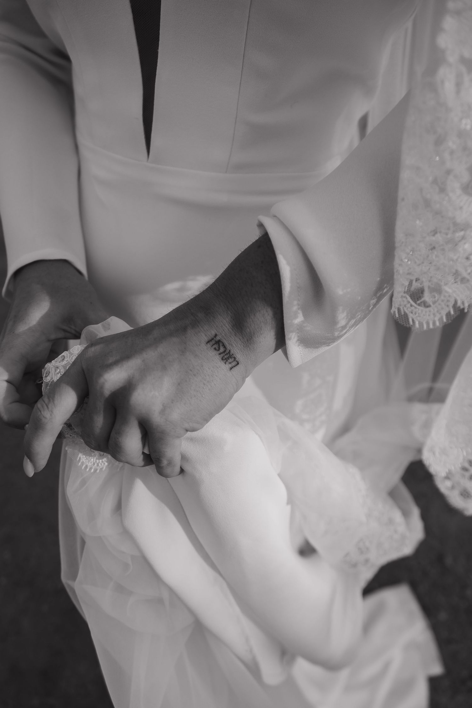 Black and white photo of bride and groom holding hands, groom's wrist tattoo visible, wedding attire detail shot.