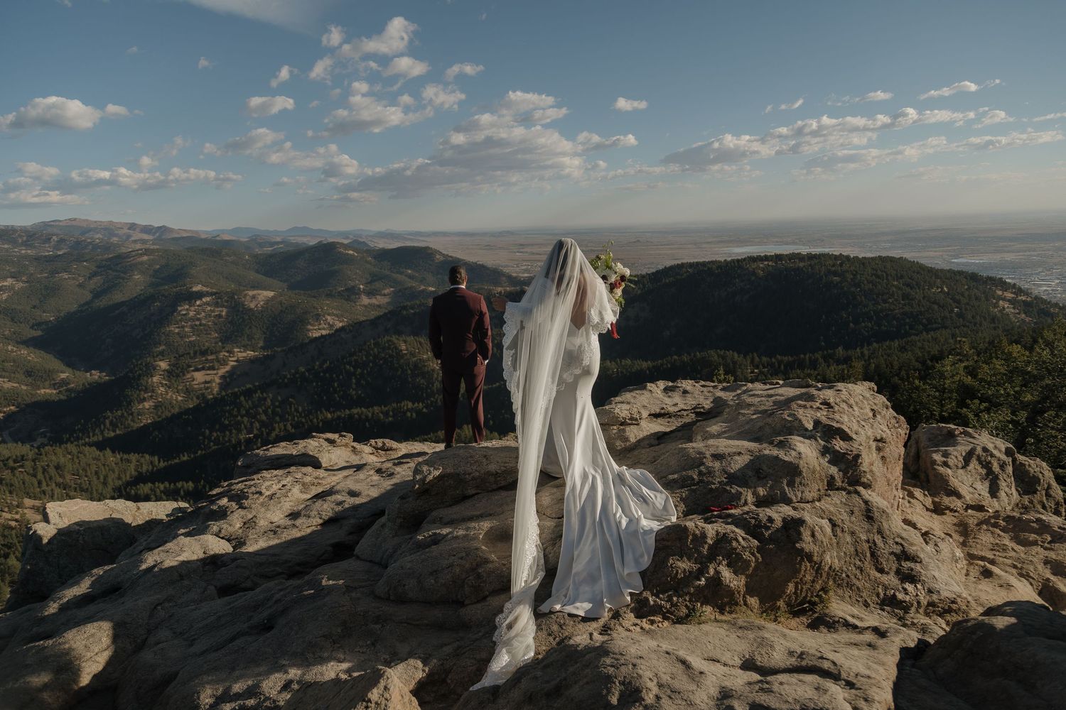 Bride in flowing white gown and long veil standing with groom overlooking breathtaking mountain valley landscape.
