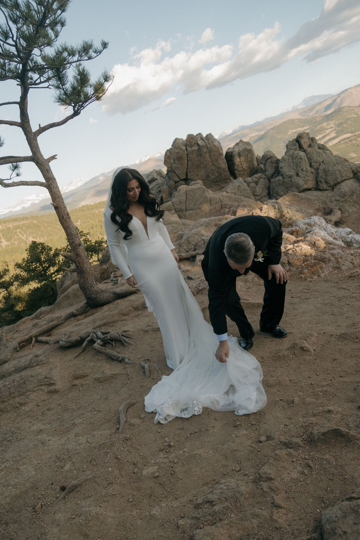 Bride in white gown with groom adjusting her train on rocky mountain overlook in Colorado.