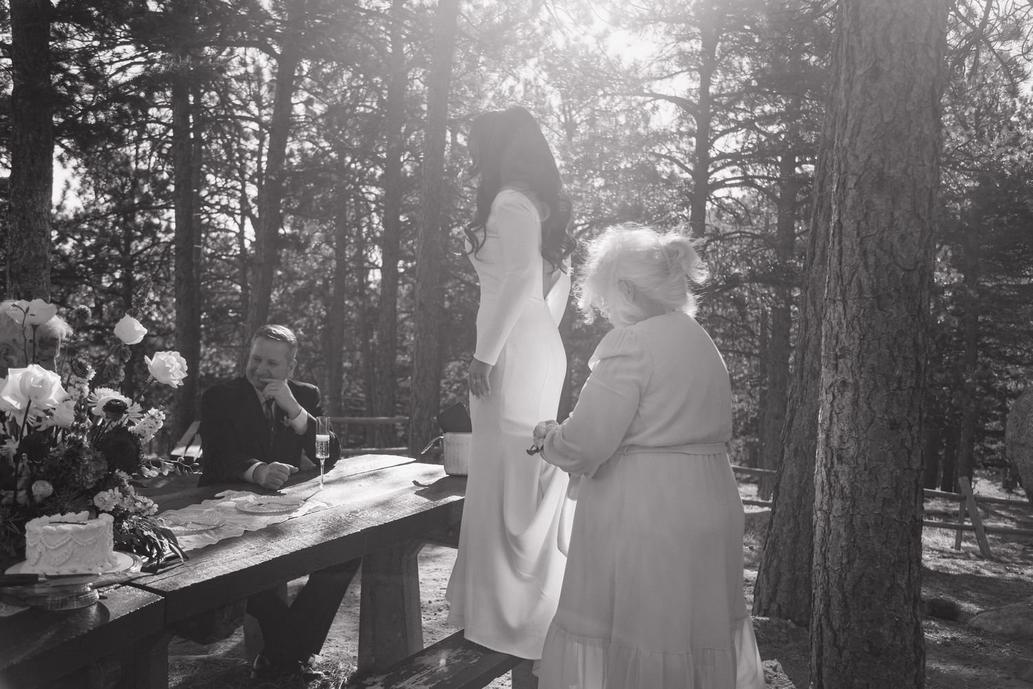 Black and white photo of bride signing marriage license at outdoor woodland table with family witnesses present.