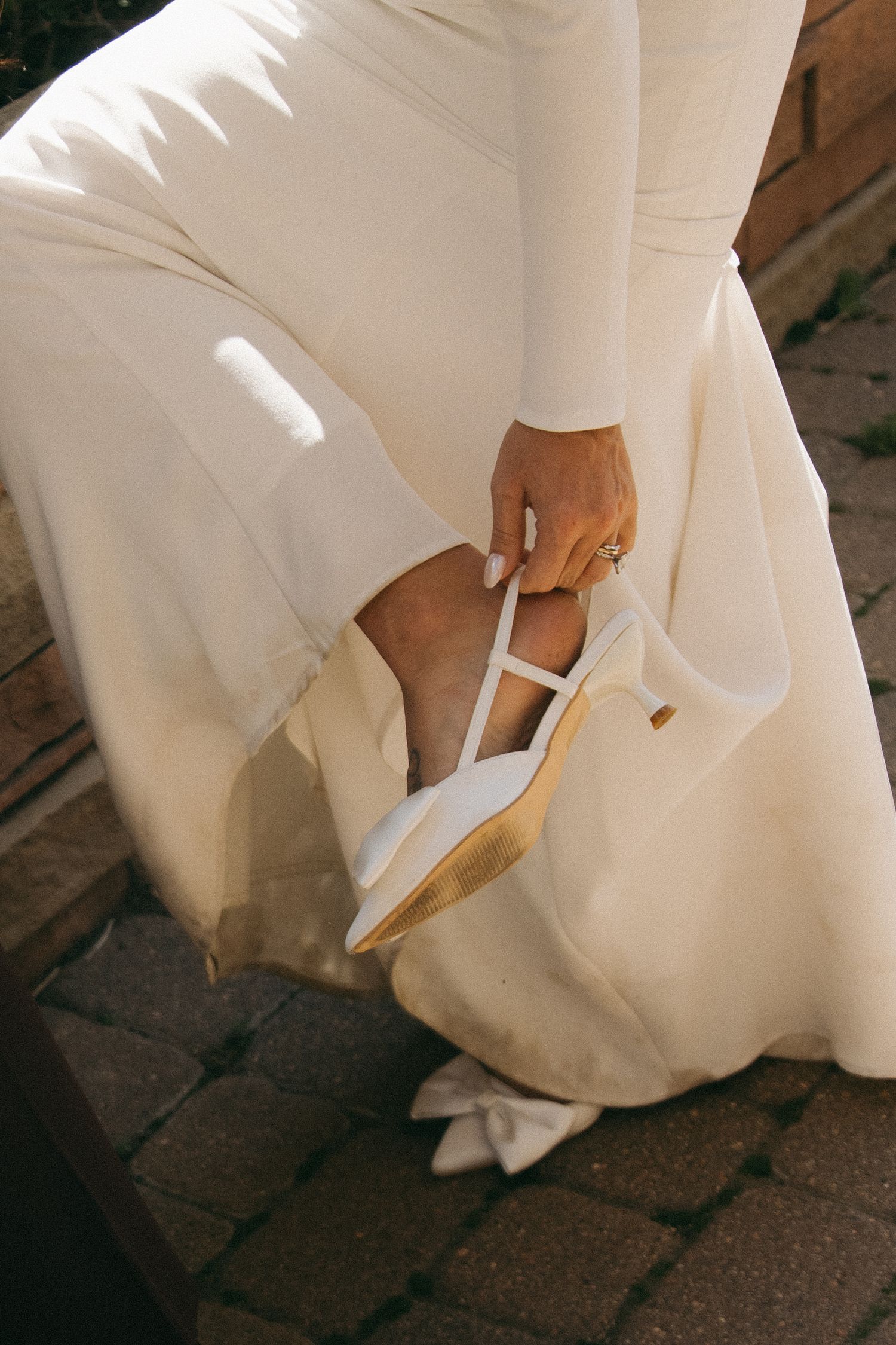 Close-up of bride in white gown adjusting white T-strap wedding heels on brick pavement.