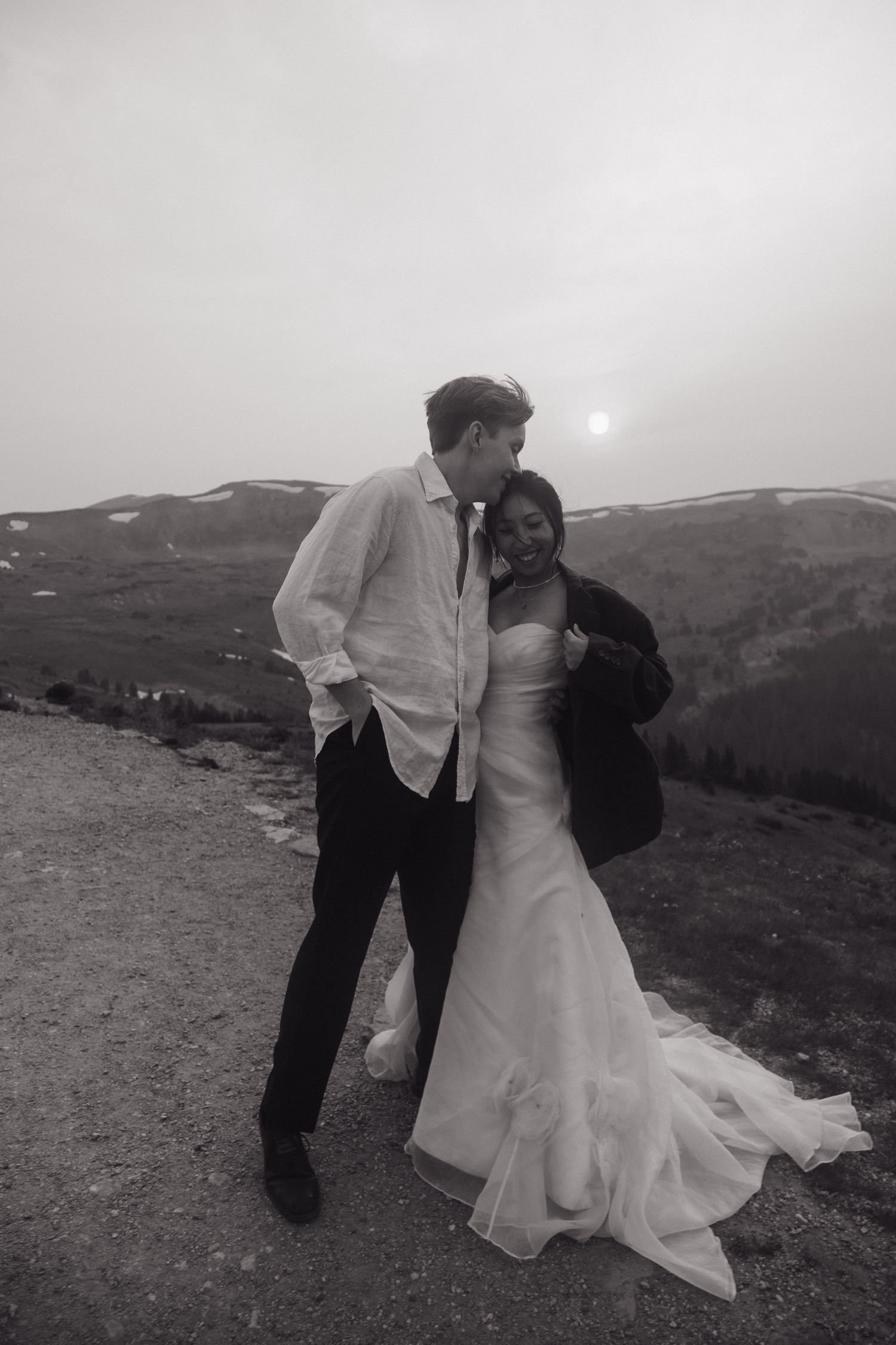 Black and white photo of laughing couple on mountain ridge at sunset, bride in gown with groom in white shirt.