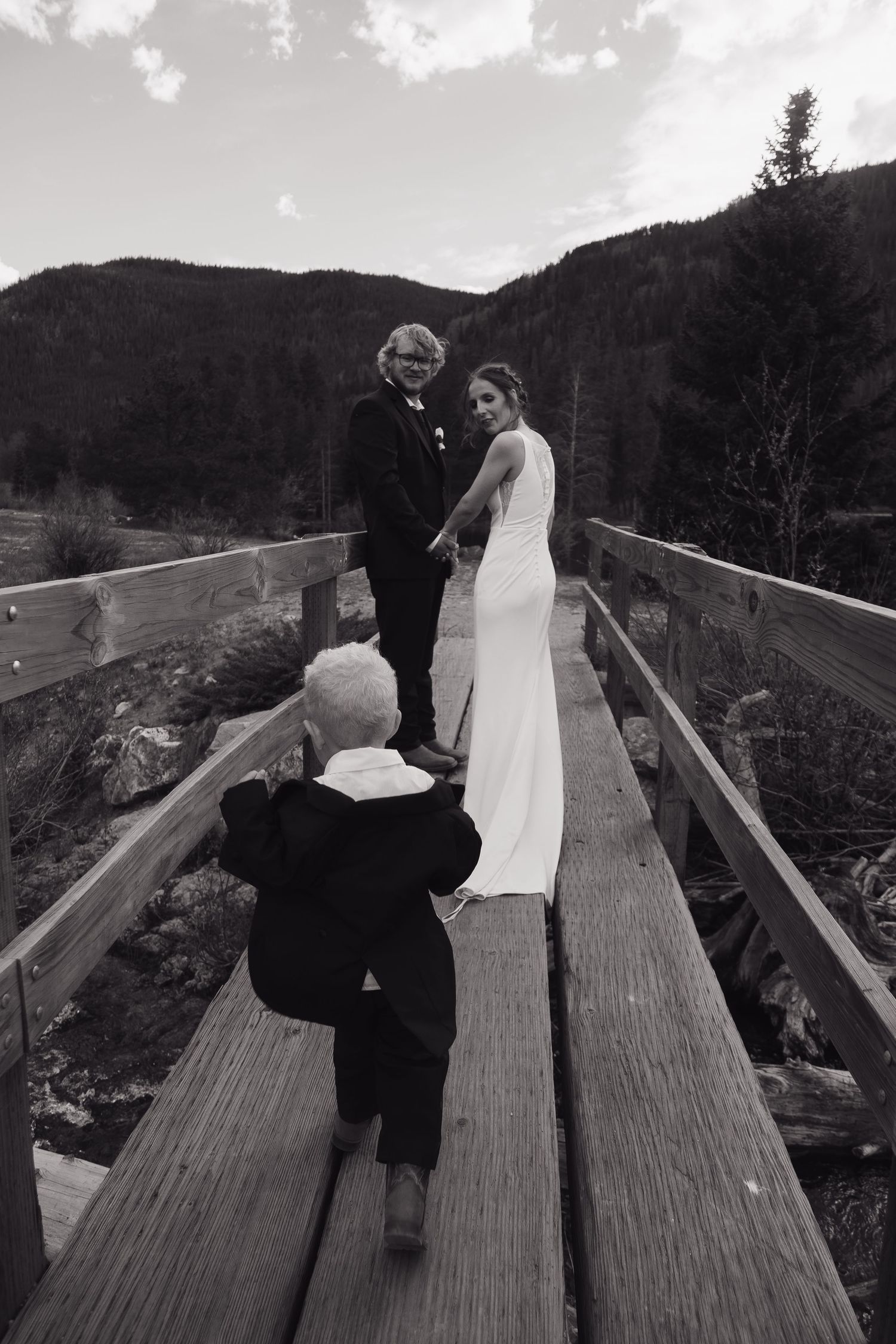 Child photographs bride and groom holding hands on rustic wooden bridge.
