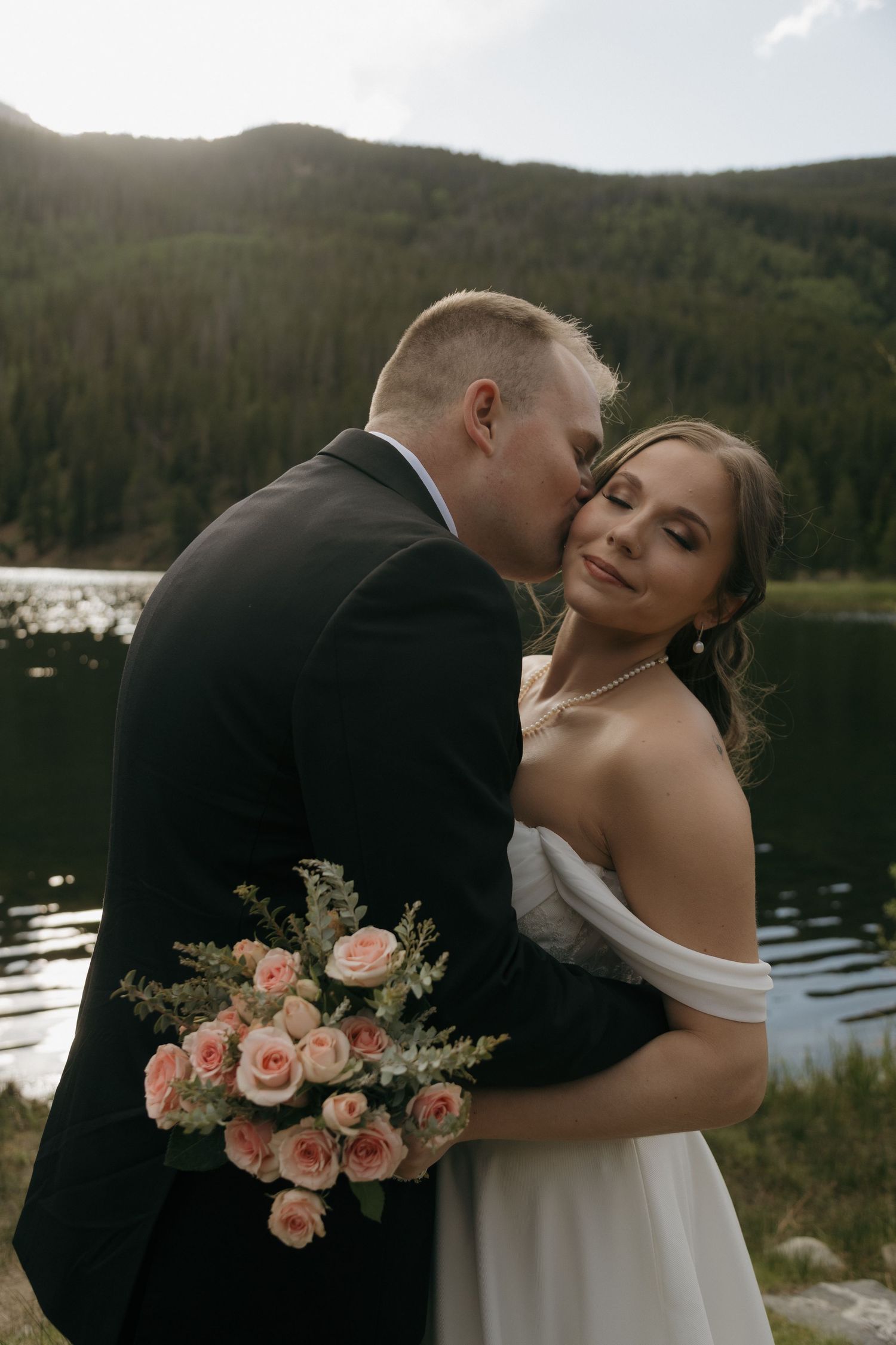 Groom in black suit kisses bride in off-shoulder white gown holding pink roses bouquet by mountain lake.