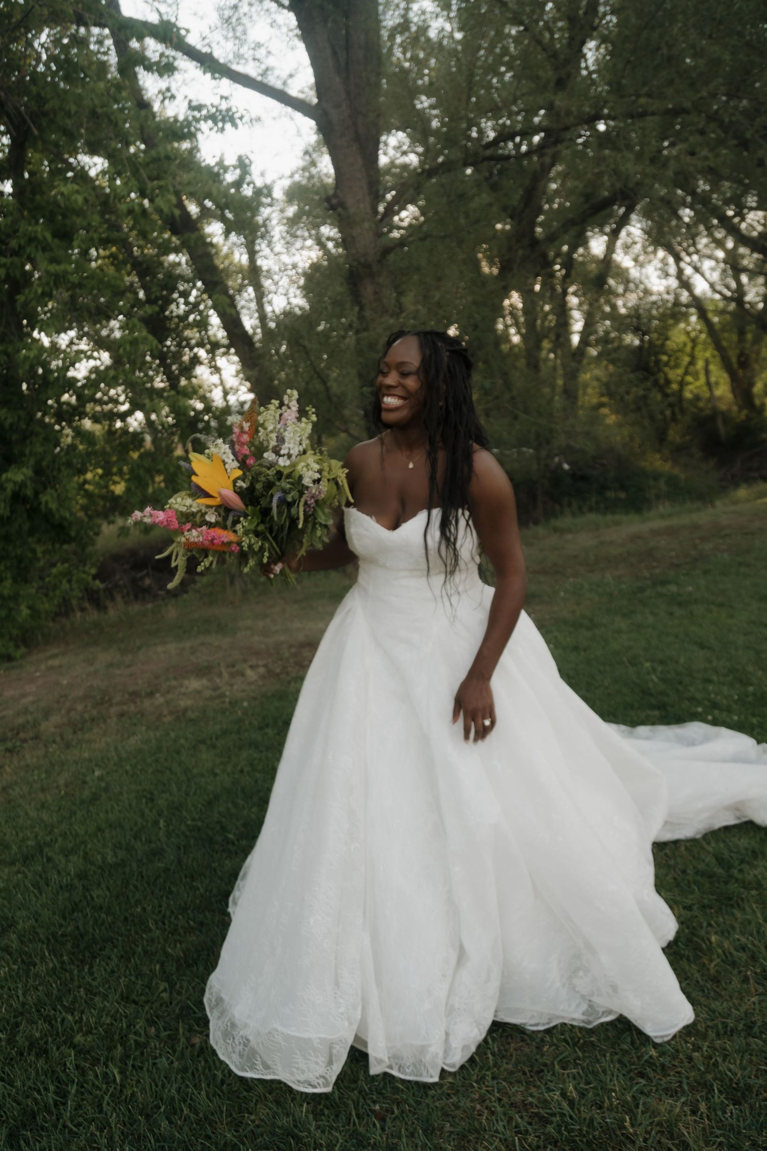 Joyful Black bride in a white ballgown laughing while holding a colorful bouquet in a lush green garden.