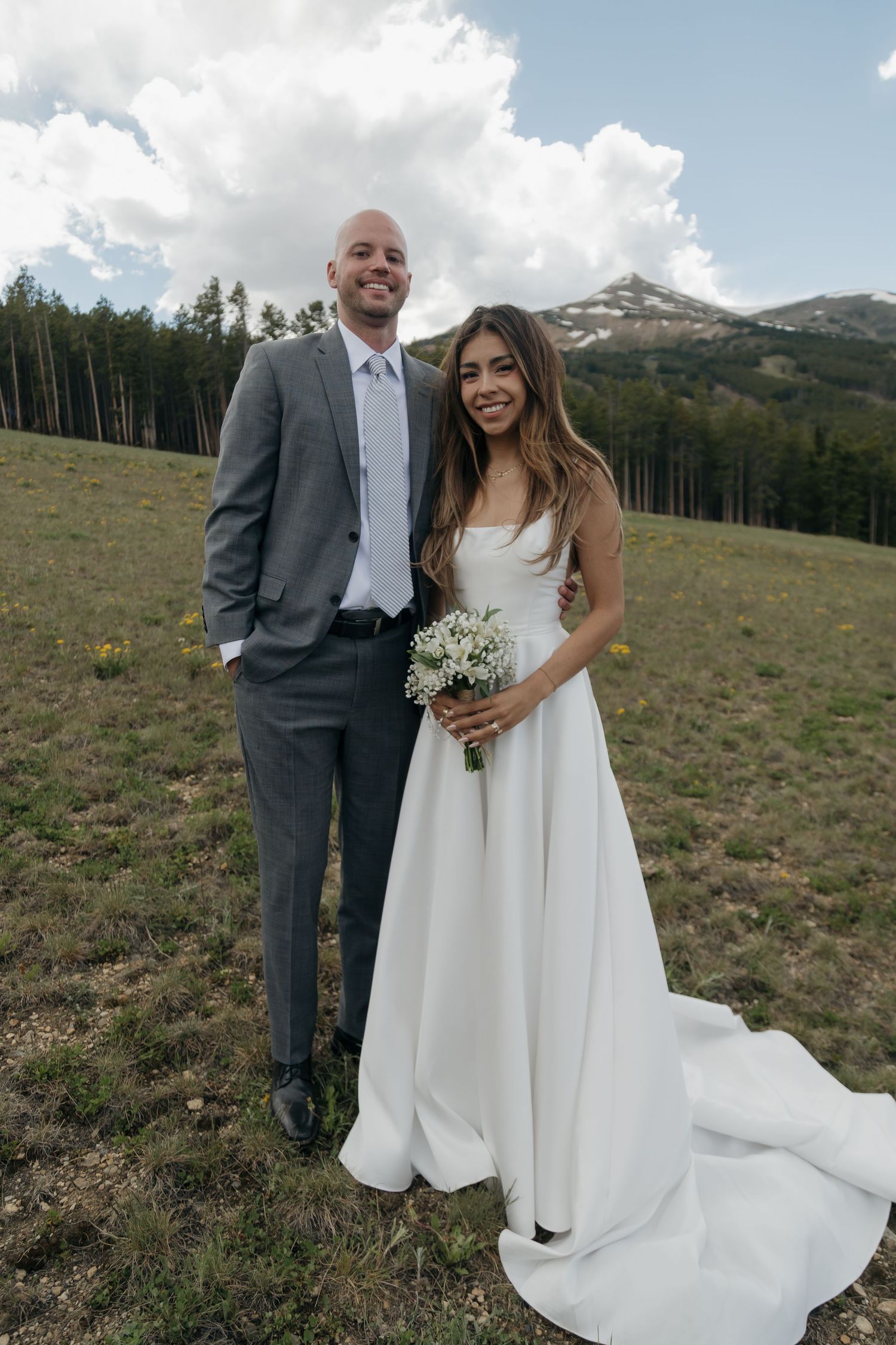 Smiling bride and groom pose together in a mountain meadow, bride holding a white bouquet on their wedding day.